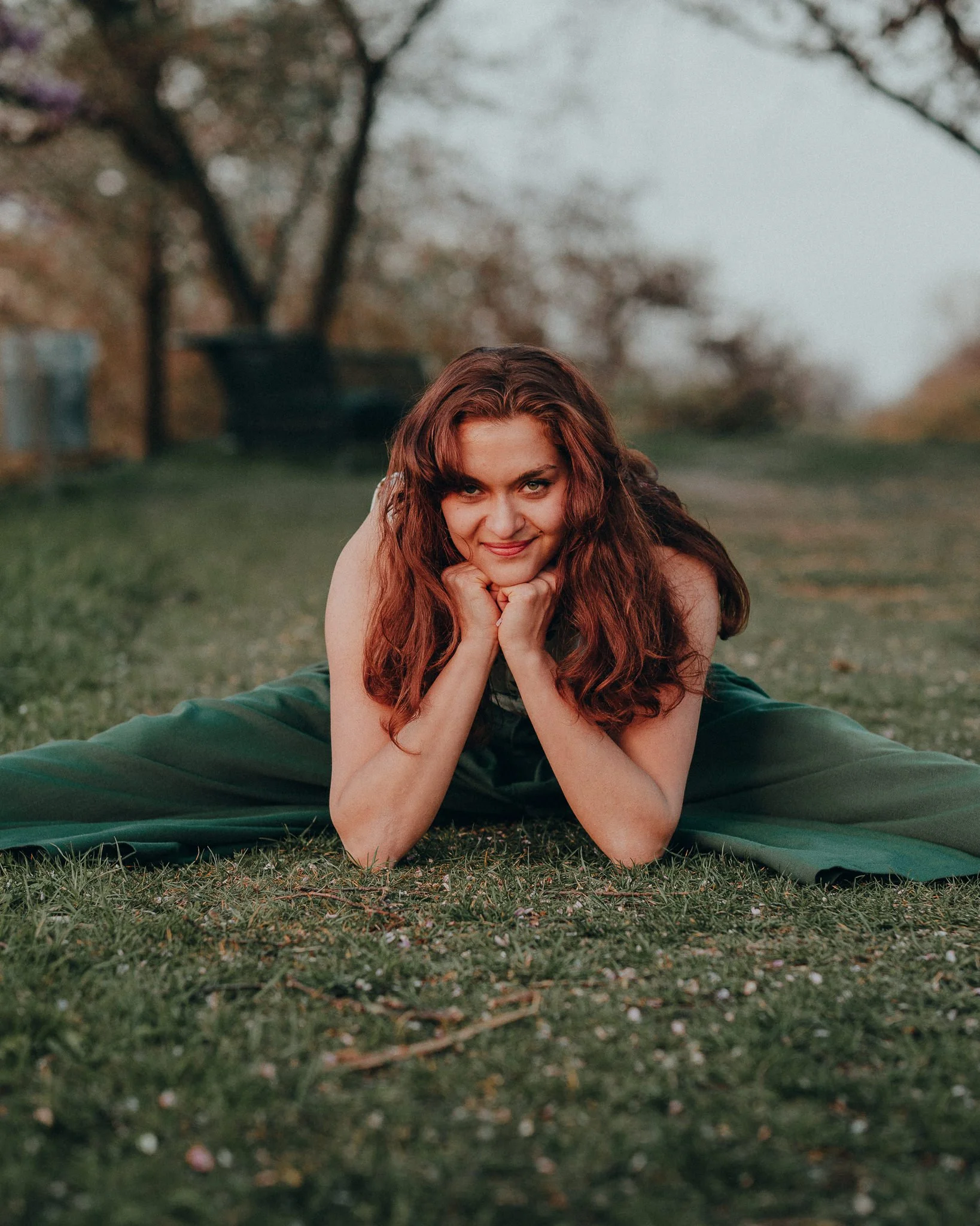 Eine junge Frau liegt auf dem Bauch im Gras, stützt ihr Kinn mit den Händen und lächelt in die Kamera, im Hintergrund Bäume und ein Himmel bei Sonnenuntergang.