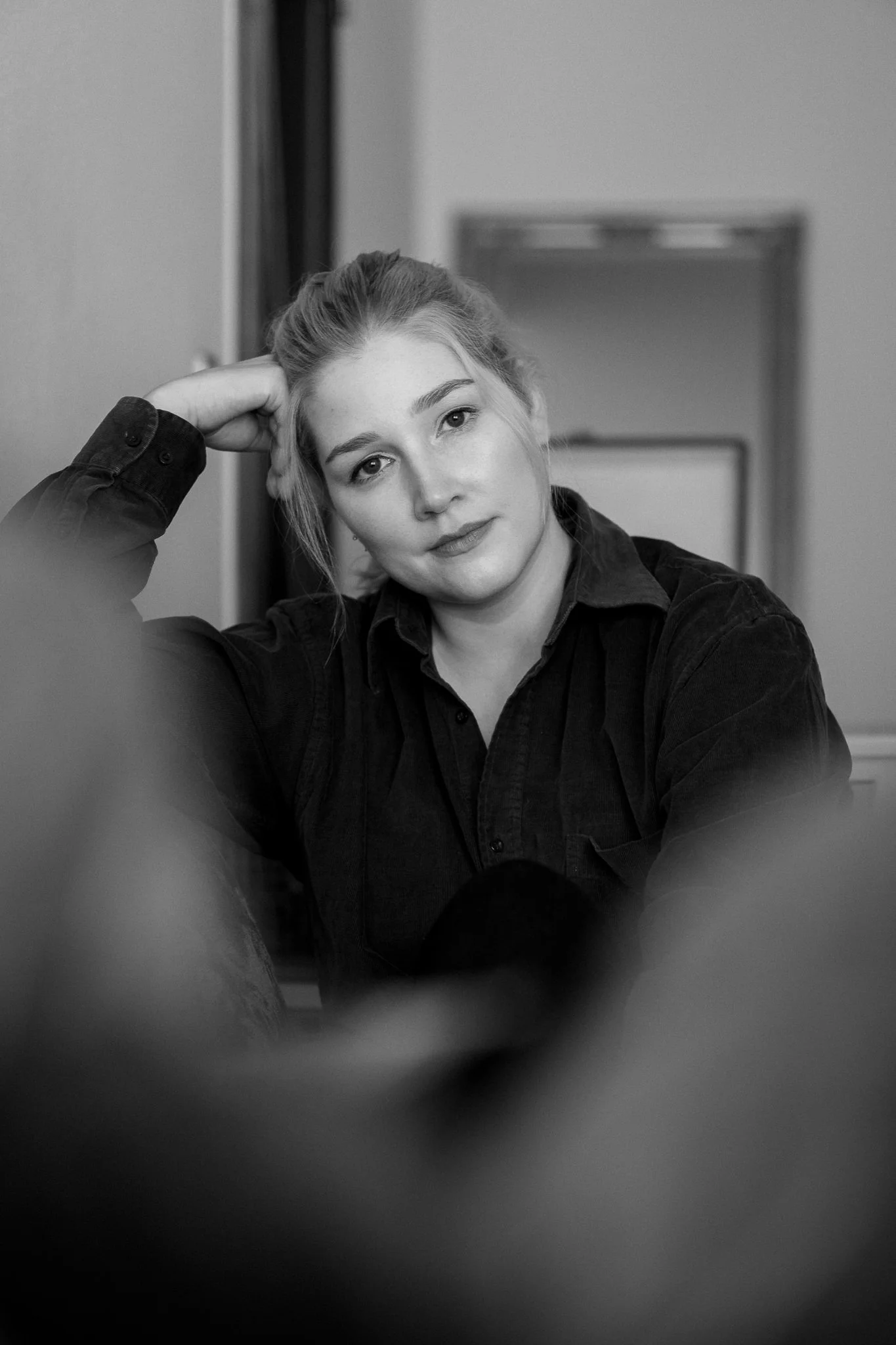 Schwarz-weiß-Fotografie eines jungen Mädchens mit aufgenommenem Haar, das entspannt und nachdenklich wirkt, sitzt in einem Raum mit offenem Türrahmen im Hintergrund.