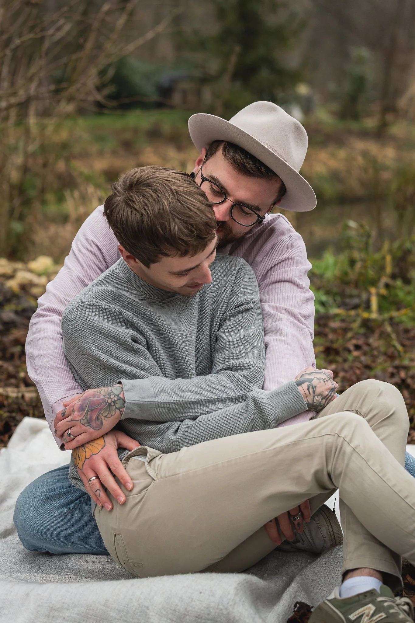 Zwei Männer umarmen sich liebevoll im Freien, im Hintergrund sind Bäume und ein Bach zu sehen.