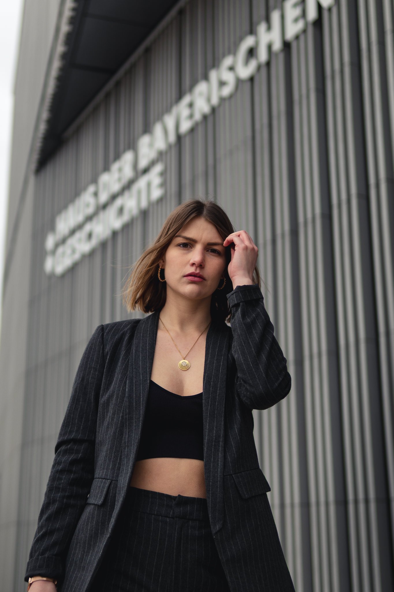 Junge Frau steht vor einem modernen Gebäude mit silberner Fassade und großen weißen Buchstaben, trägt einen schwarzen Anzug mit gestreiften Mustern, ein schwarzes Crop-Top, Goldschmuck und schaut ernst in die Kamera.