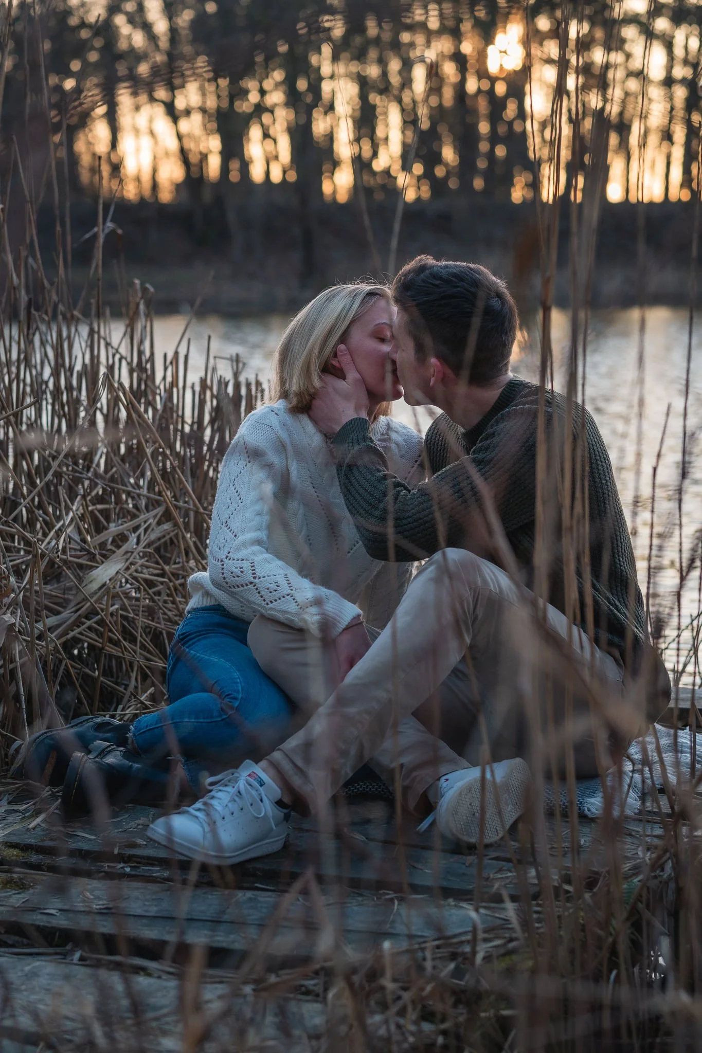 Ein Paar küsst sich am See bei Sonnenuntergang, umgeben von trockenen Schilfrohren, sitzend auf einem Holzsteg.