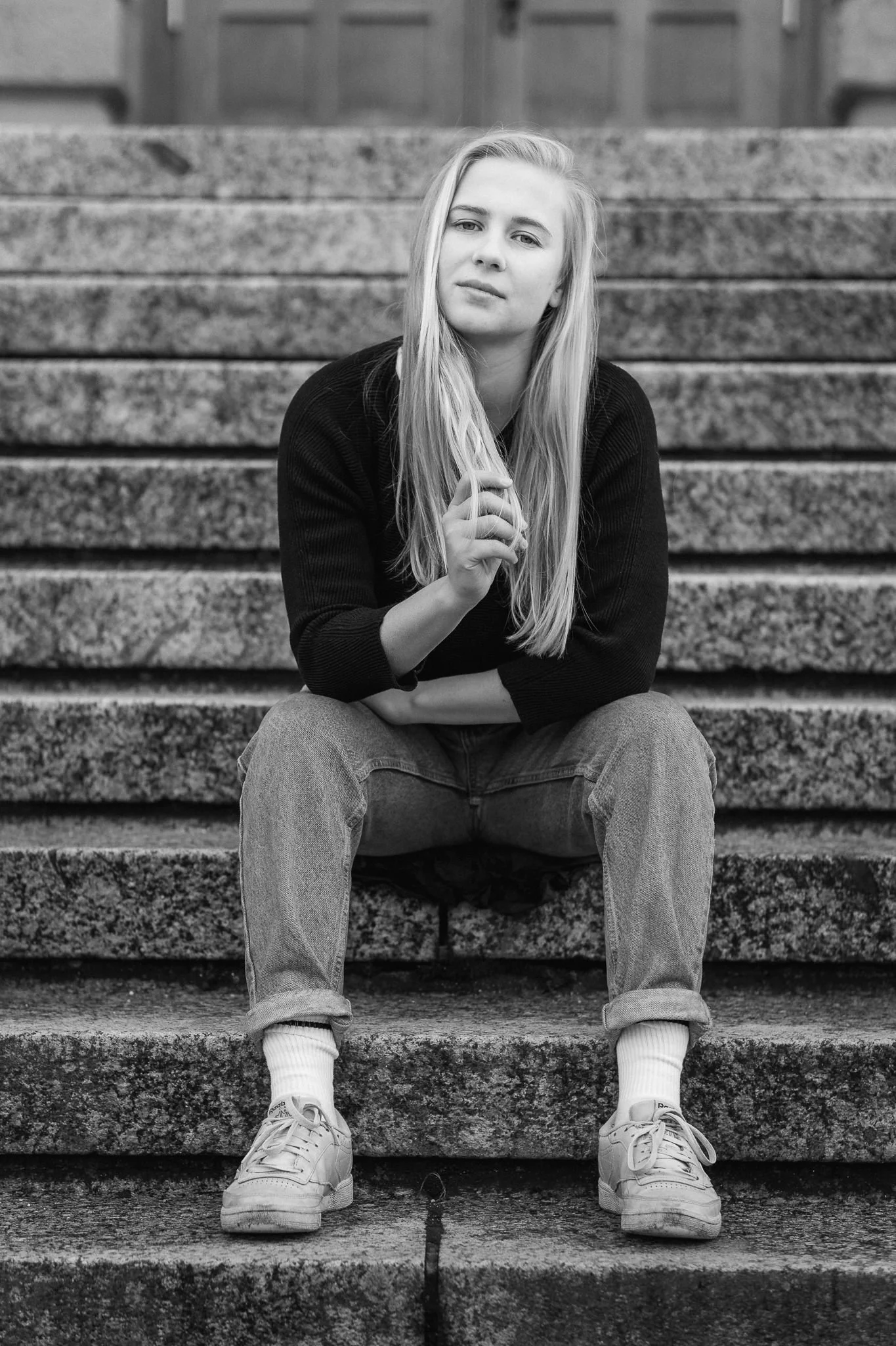 Junge Frau mit langen Haaren sitzt auf Steintreppen, trägt Sneakers, Jeans und einen Pullover, blickt in die Kamera, im Schwarz-Weiß-Foto.