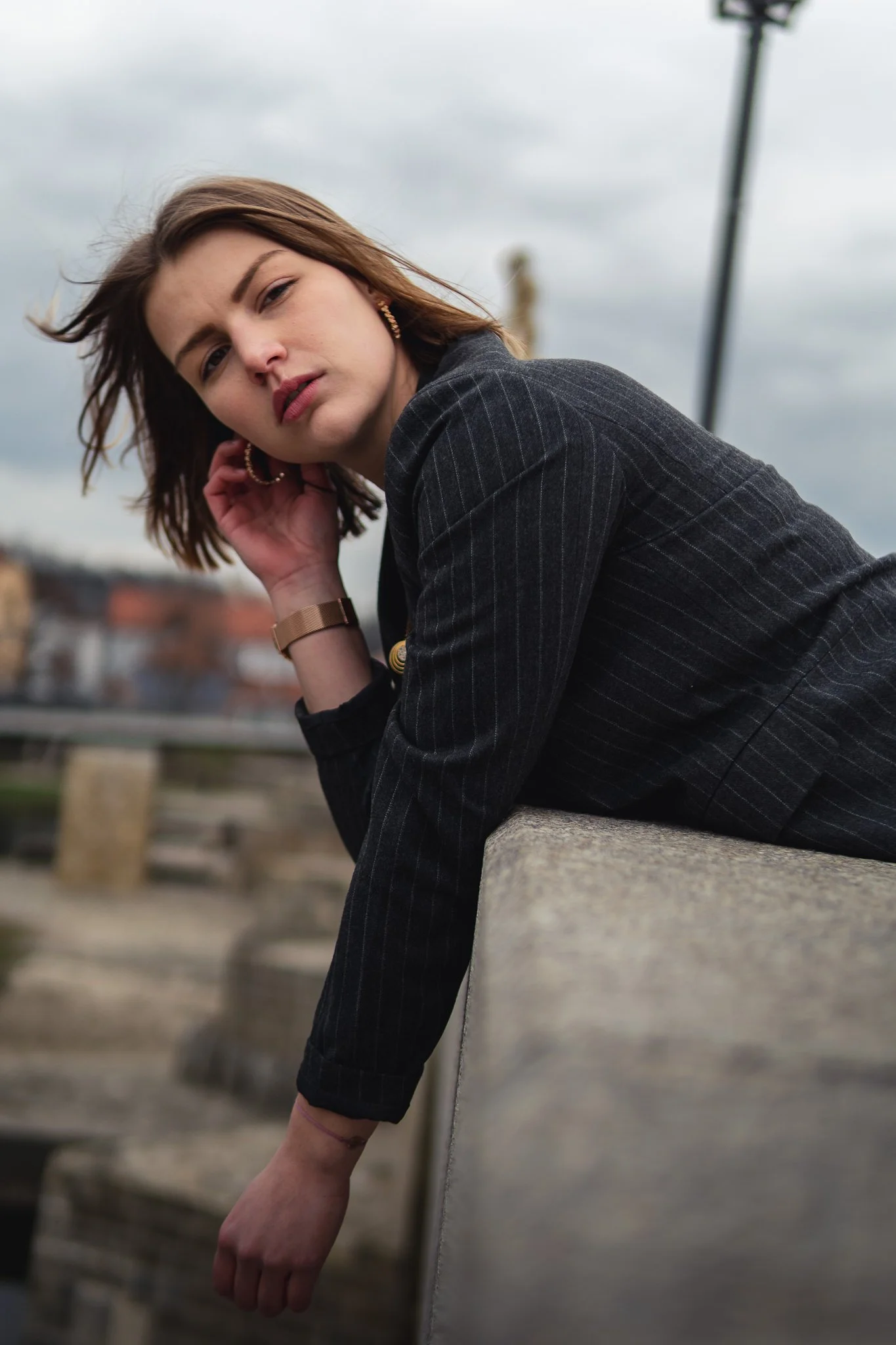 Junge Frau in dunklem Anzug lehnt an einer Betonmauer im Freien bei bewölktem Himmel.