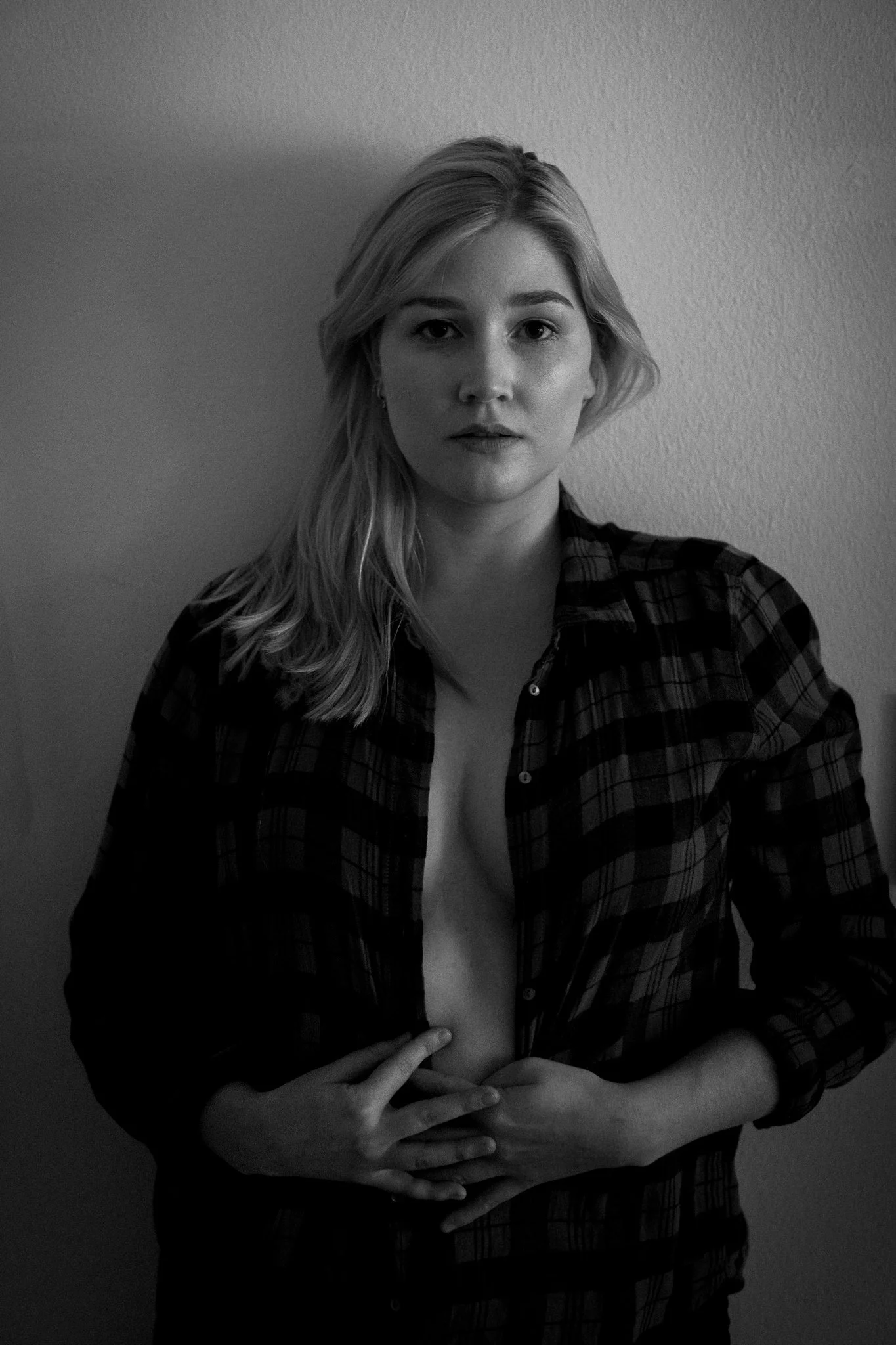 Schwarz-Weiß-Foto einer jungen Frau mit offenen, halblangen blonden Haaren, die eine offen-zu-Klappe kariertes Hemd trägt, das sie teilweise offen hält, und auf eine neutrale Wand blickt.