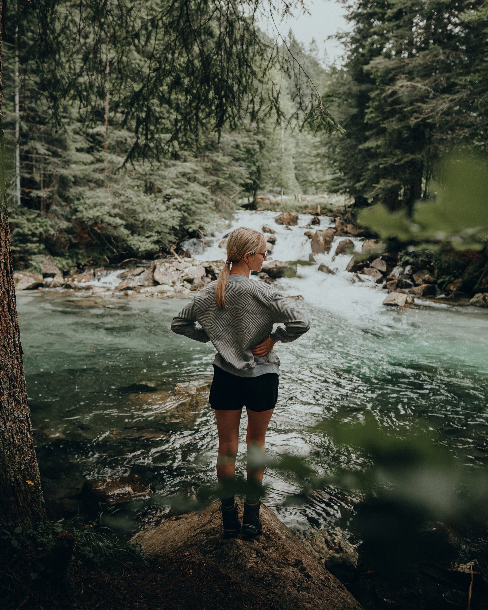 Eine Frau steht auf einem Felsen im Wasser eines Flusses, umgeben von dichten Wald mit hohen Bäumen, während sie die Natur betrachtet.
