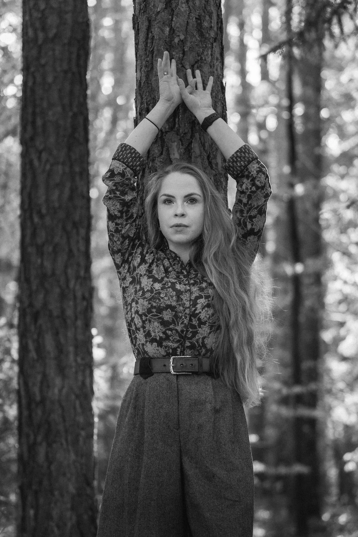Schwarz-weiß Foto einer Frau mit langen Haaren, die in einem Wald gegen einen Baum hält, mit den Händen hoch oben an den Baumstamm.