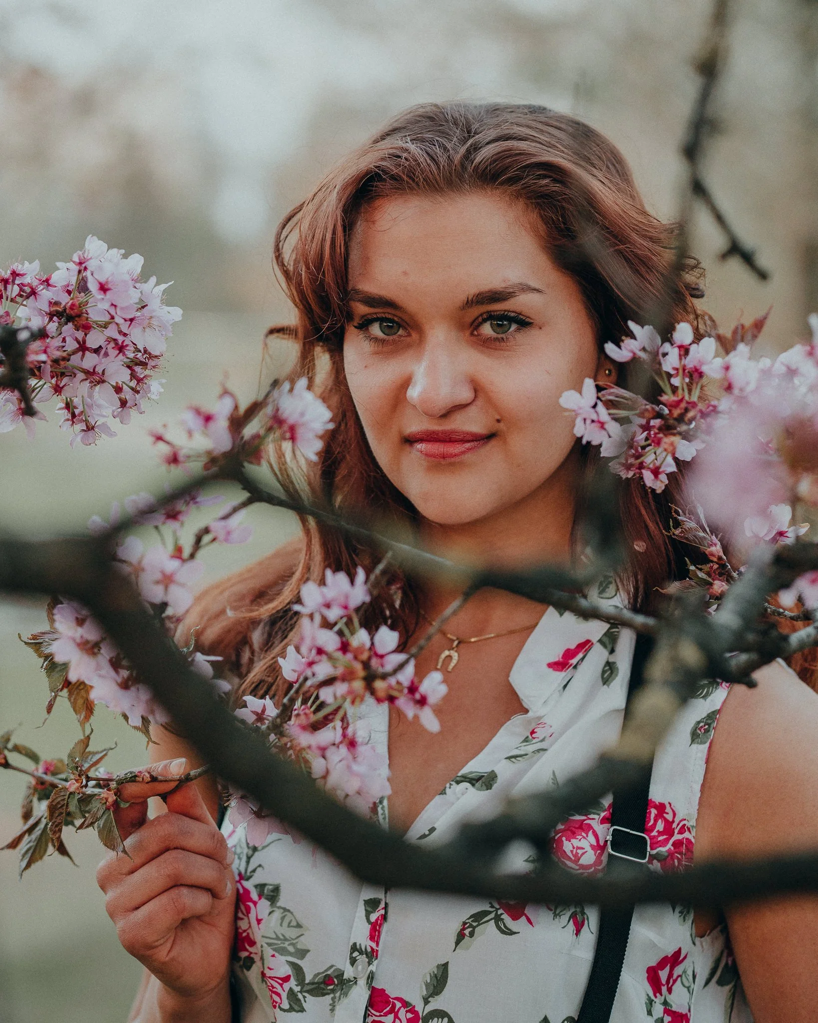 Eine junge Frau mit braunen, welligen Haaren, die vor blühenden rosa-weißen Kirschblüten steht.