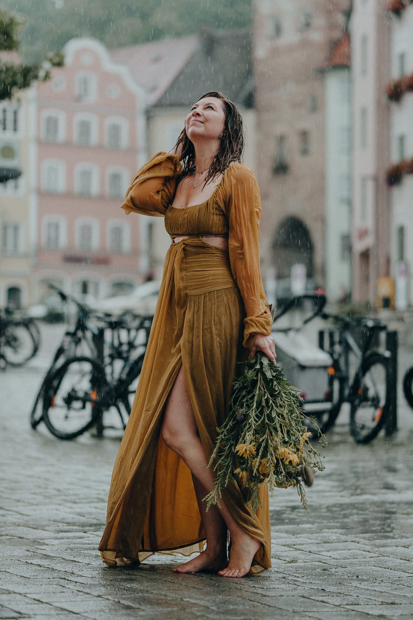 Eine Frau in einem goldbraunen Kleid steht barfuß auf einem gepflasterten Platz im Regen, hält einen Blumenstrauß und lächelt, während bunte Gebäude im Hintergrund sichtbar sind.