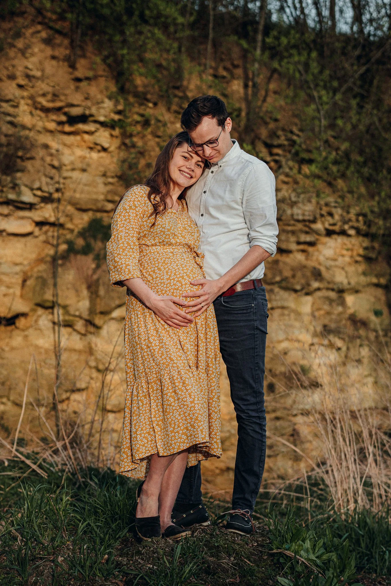 Ein glückliches Paar im Freien, das eine schwangere Frau liebevoll hält. Im Hintergrund eine Felsenwand und Natur.