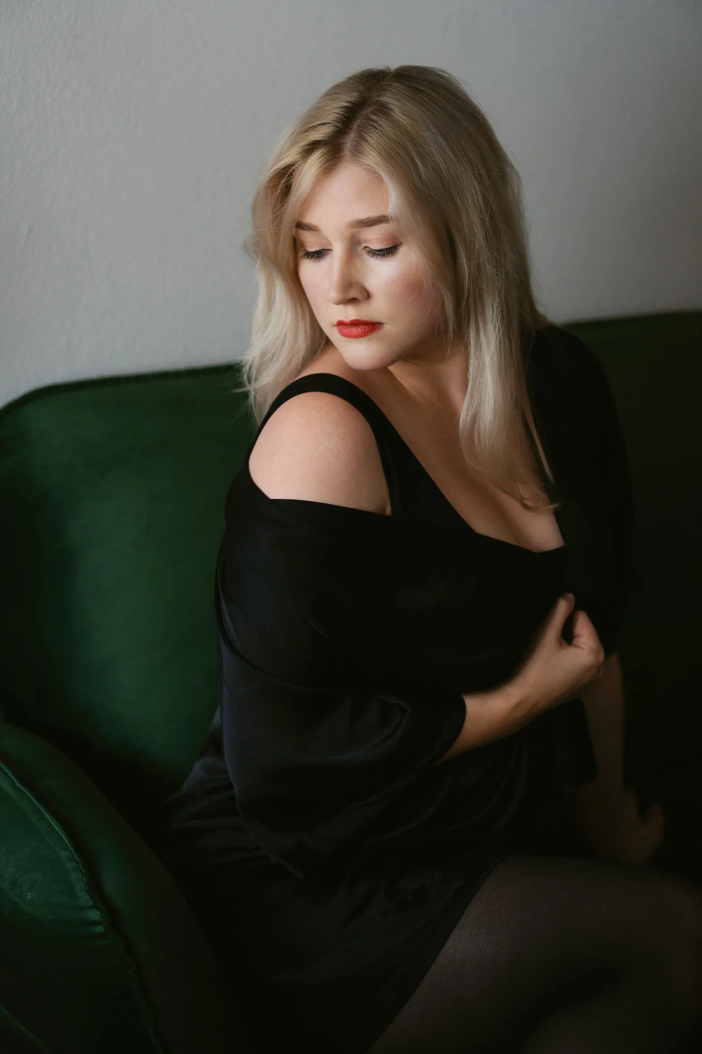 Junge Frau mit blonden Haaren sitzt auf einem grünen Sofa, trägt ein schwarzes Kleid mit offenem Schulterdesign, schaut nach unten und hält eine Hand an die Brust.