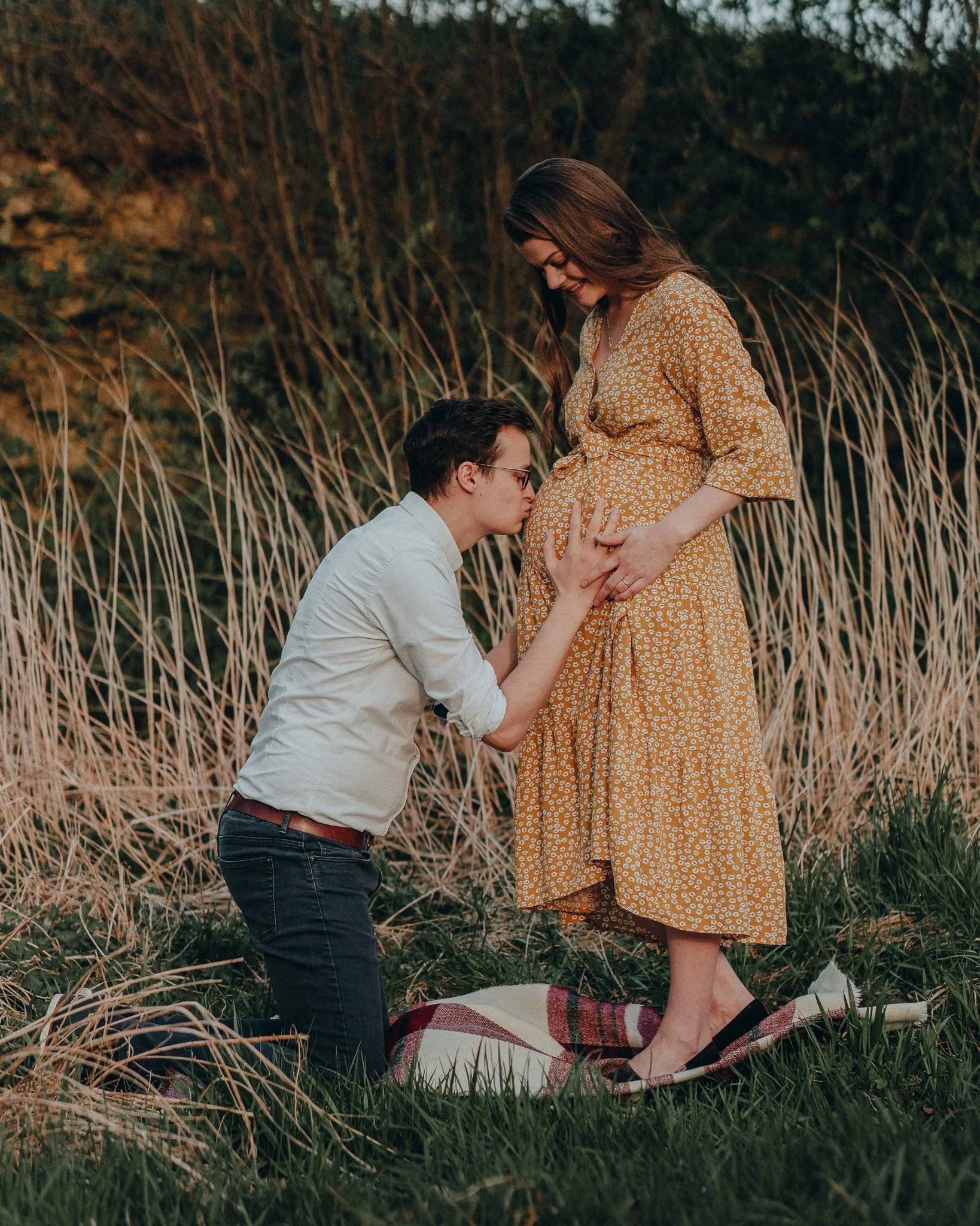 Ein Mann kniet vor einer schwangeren Frau auf einem Picknickdecke im Freien, sie hat ein Kleid mit Blumenmuster. Der Mann küsst ihren Bauch, beide lächeln. Es ist Abendlicht.
