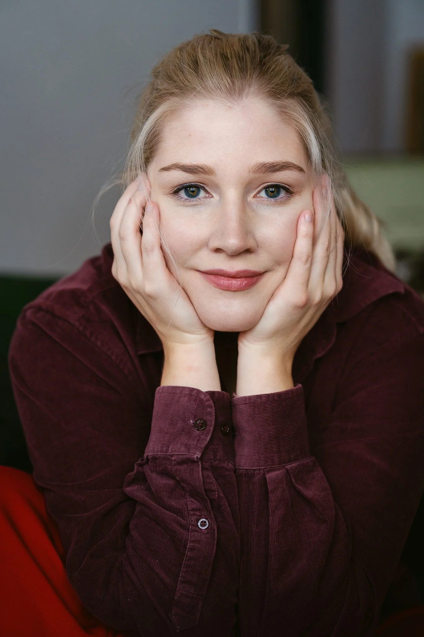 Nahaufnahme einer lächelnden Frau mit blonden Haaren, die ihre Wangen mit den Händen stützt, in einem gemütlichen Raum.