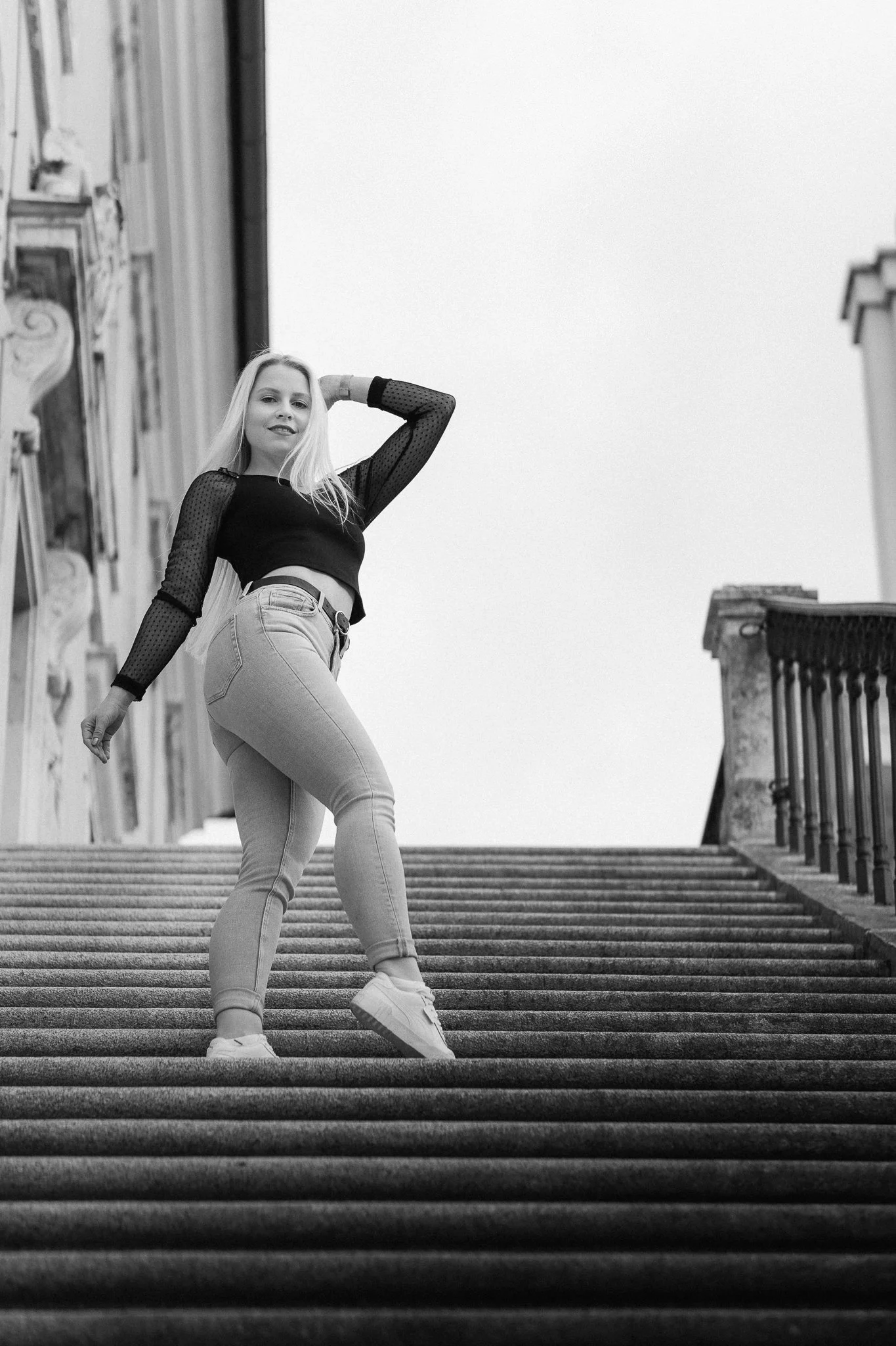Eine junge Frau posiert auf einer Außentreppe, trägt Jeans, einen schwarzen Oberteil mit transparenten, gepunkteten Ärmeln, und weiße Sneakers, im Schwarz-Weiß-Format aufgenommen im Stadtumfeld.