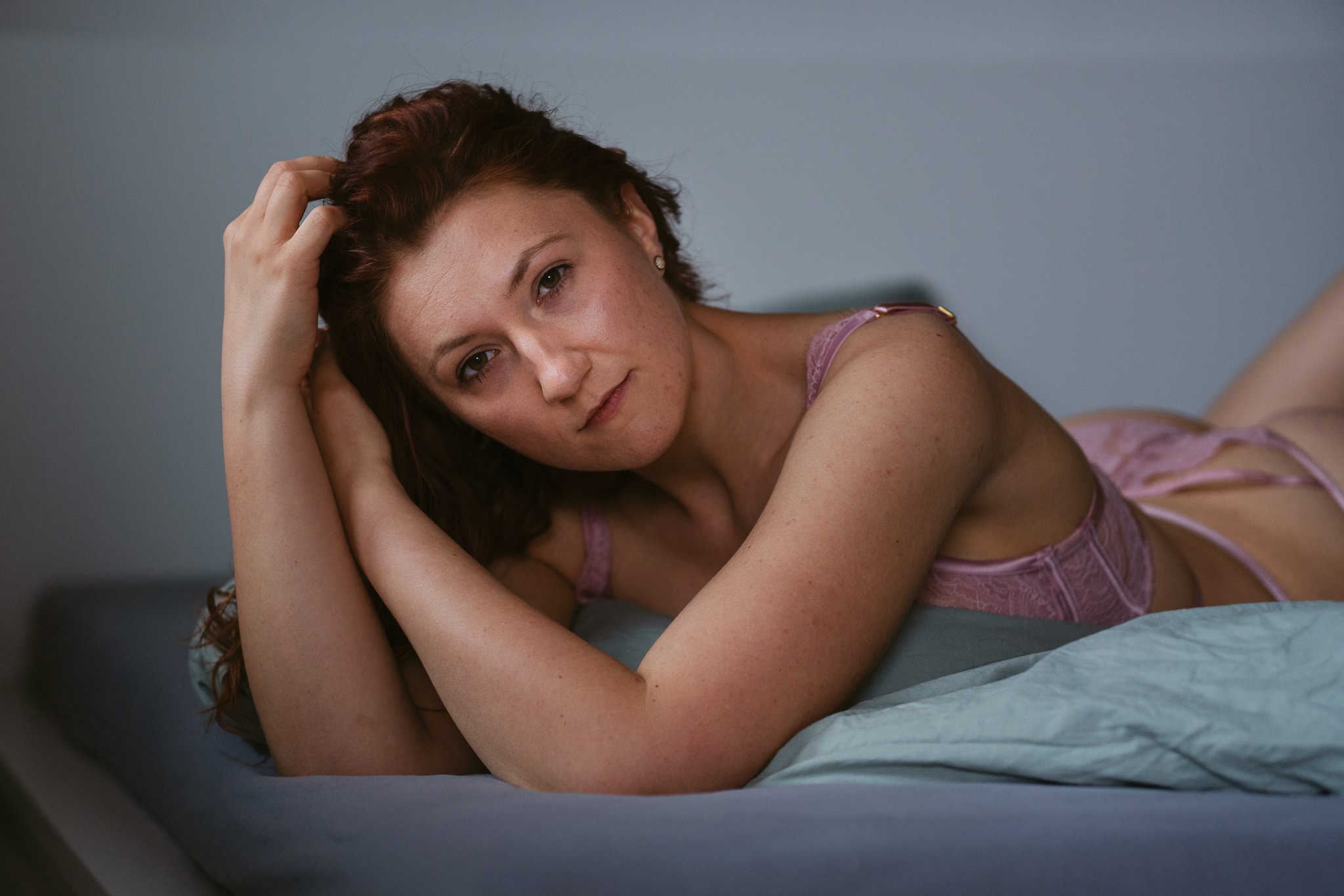 Junge Frau liegt auf einem Bett, schaut entspannt in die Kamera, trägt lila Spitze Unterwäsche, mit braunen lockigen Haaren und natürlichem Make-up.