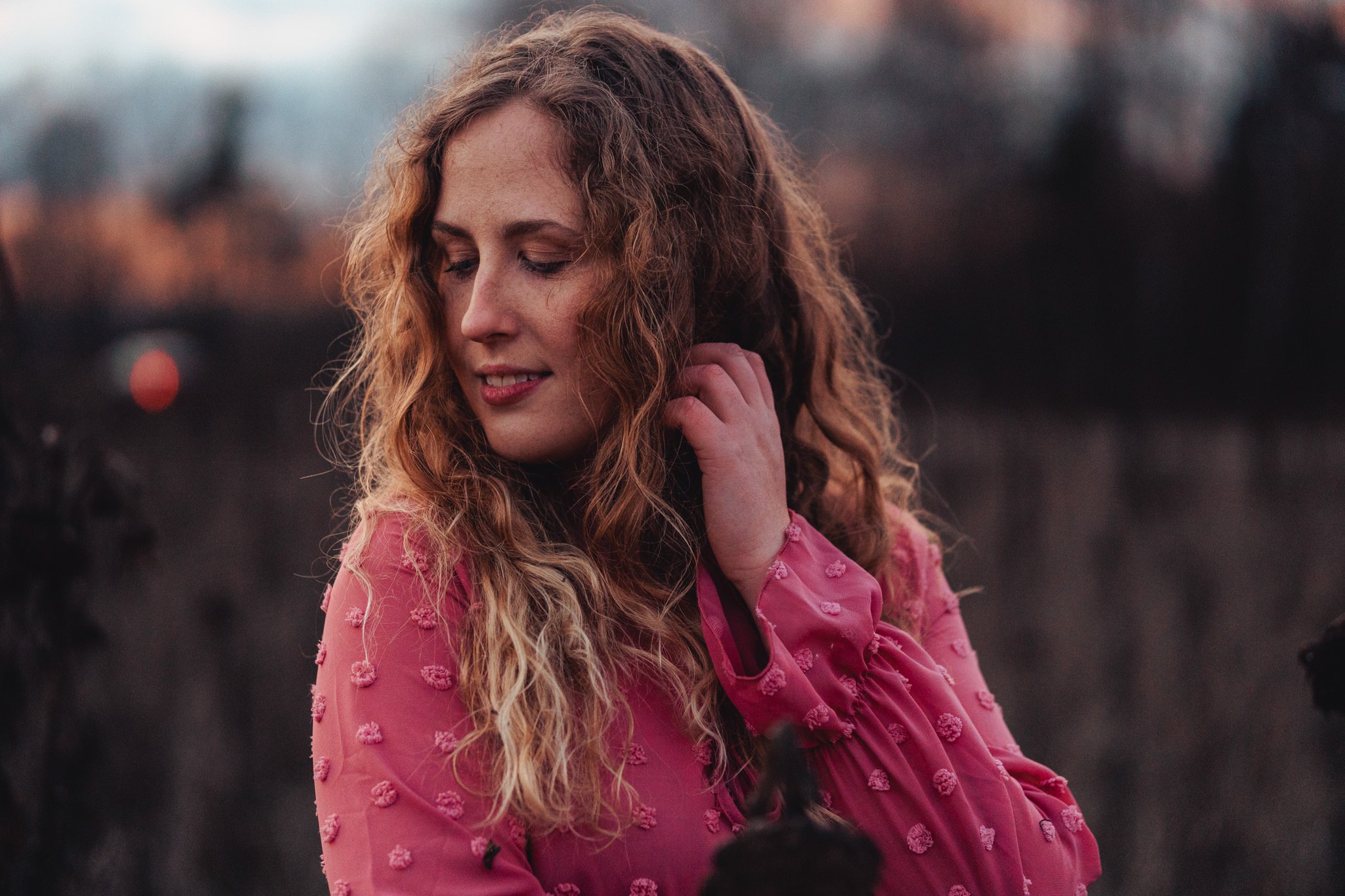 Eine junge Frau mit lockigem, roten Haar trägt ein pinkes Kleid mit kleinen Ballonmuster. Sie steht im Freien in einer dämmernden Umgebung, schaut nach unten und berührt vorsichtig ihr Ohr.