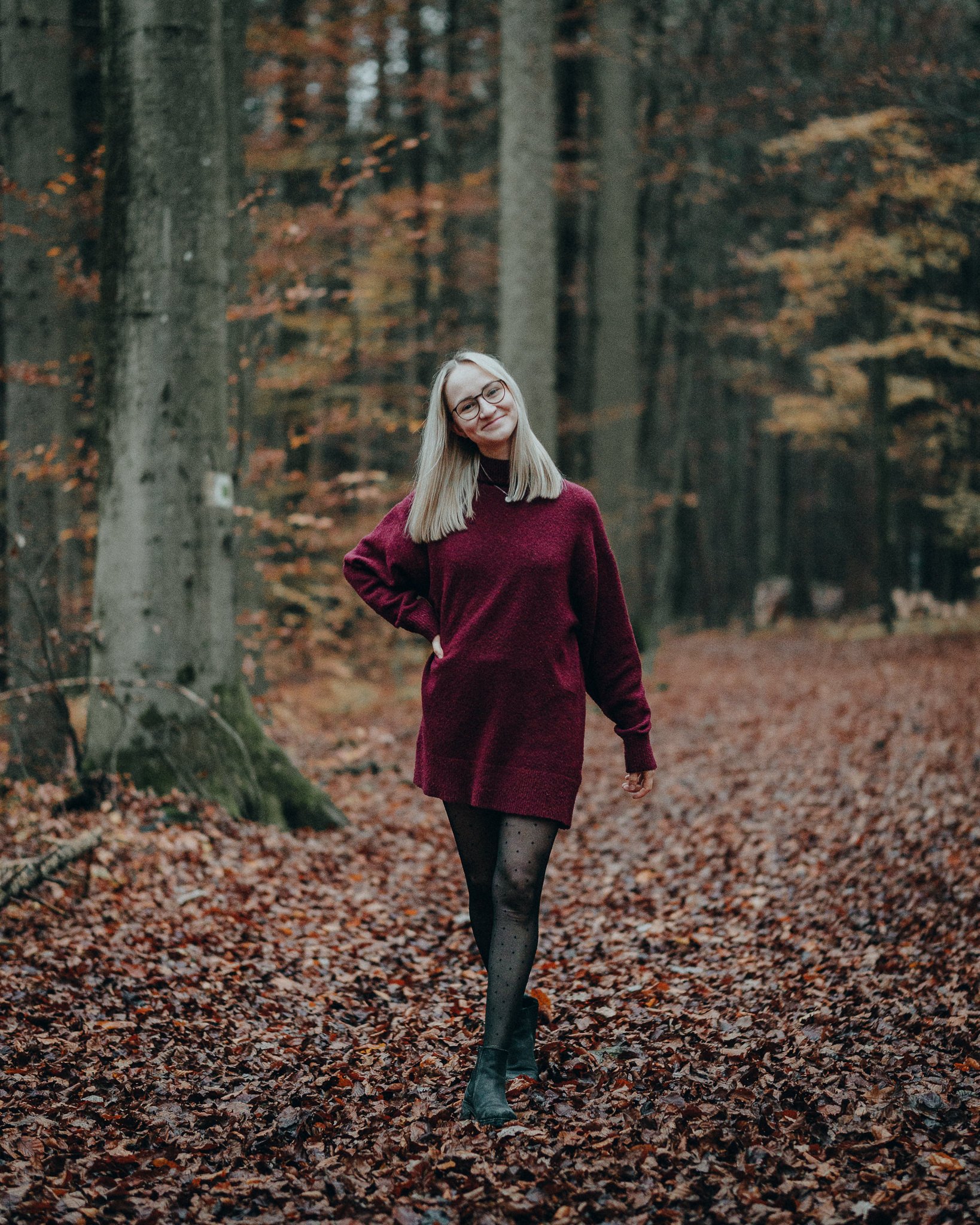 Junge Frau geht durch einen herbstlichen Wald auf einem mit fallen Blättern bedeckten Weg und trägt einen roten Pullover, schwarze Strumpfhosen mit Punkten, schwarze Stiefel und trägt Brille.