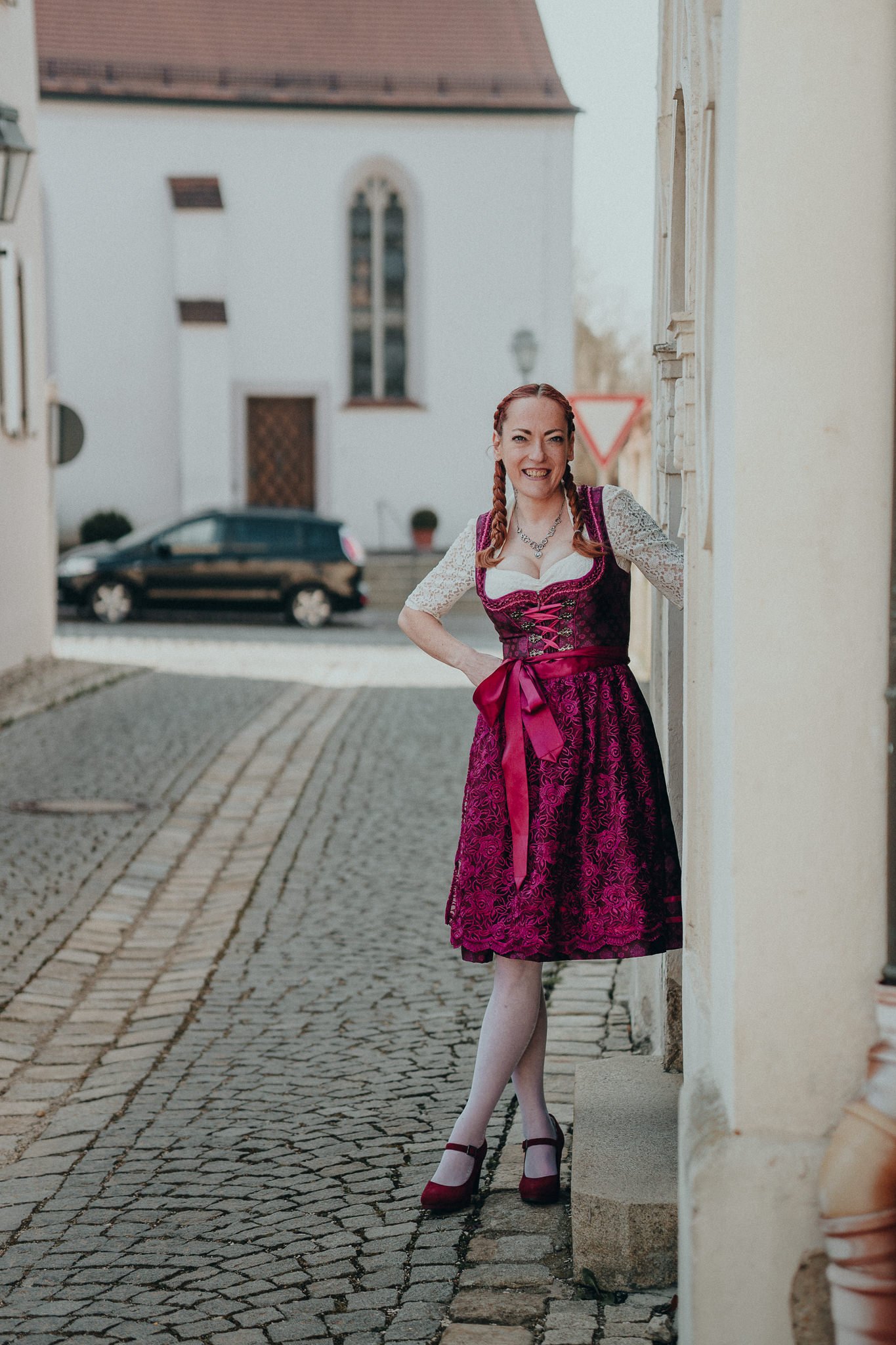 Frau in traditioneller bayerischer Tracht mit rotem Kleid, weißer Bluse und Flechtzopf, die an einer Gebäudewand in einer gepflasterten Straße steht.