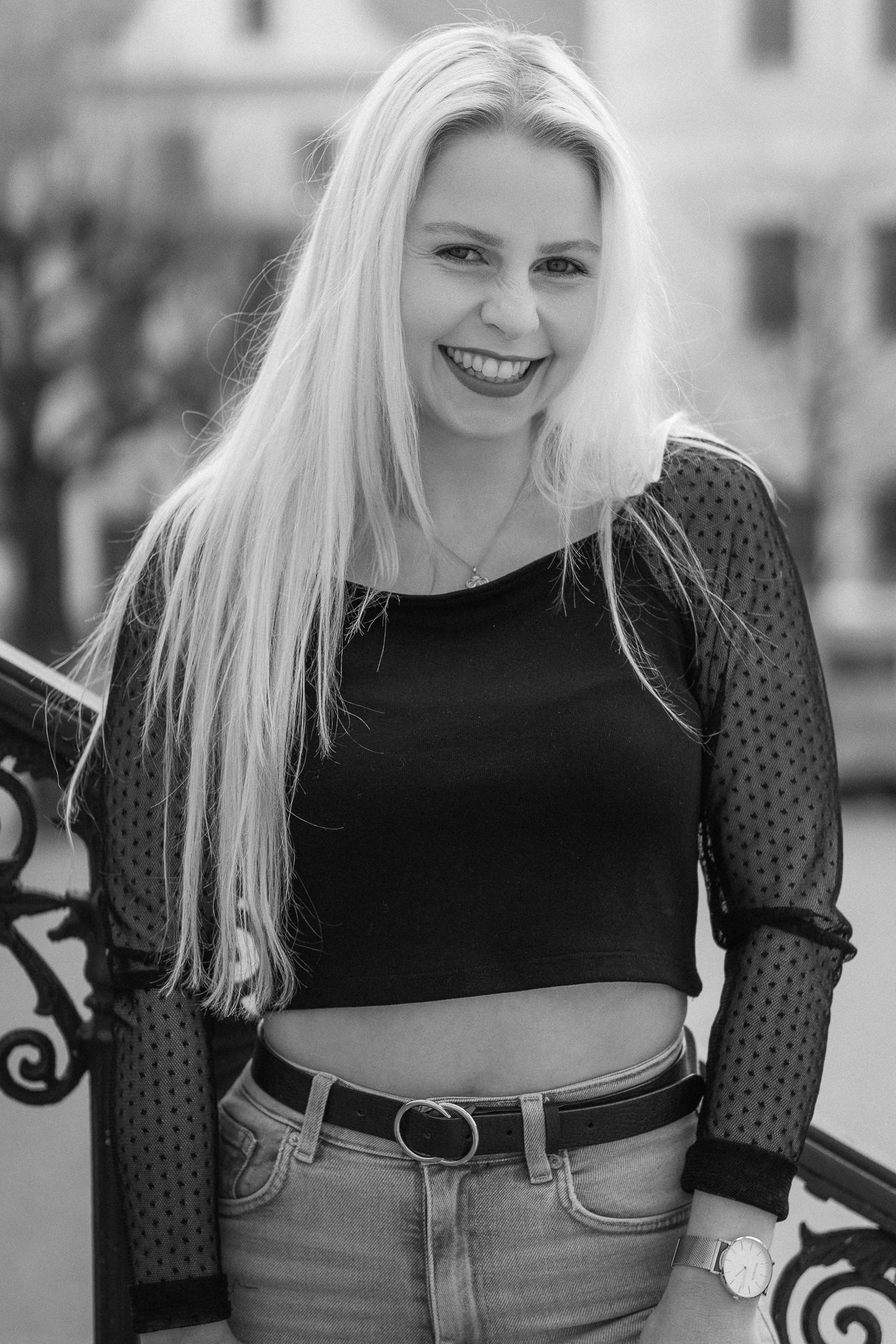 Lächelnde junge Frau mit langen blonden Haaren, tragen schwarzes Top mit transparenten, gepunkteten Ärmeln, Jeans und Uhr, steht im Freien mit unscharbem Hintergrund.