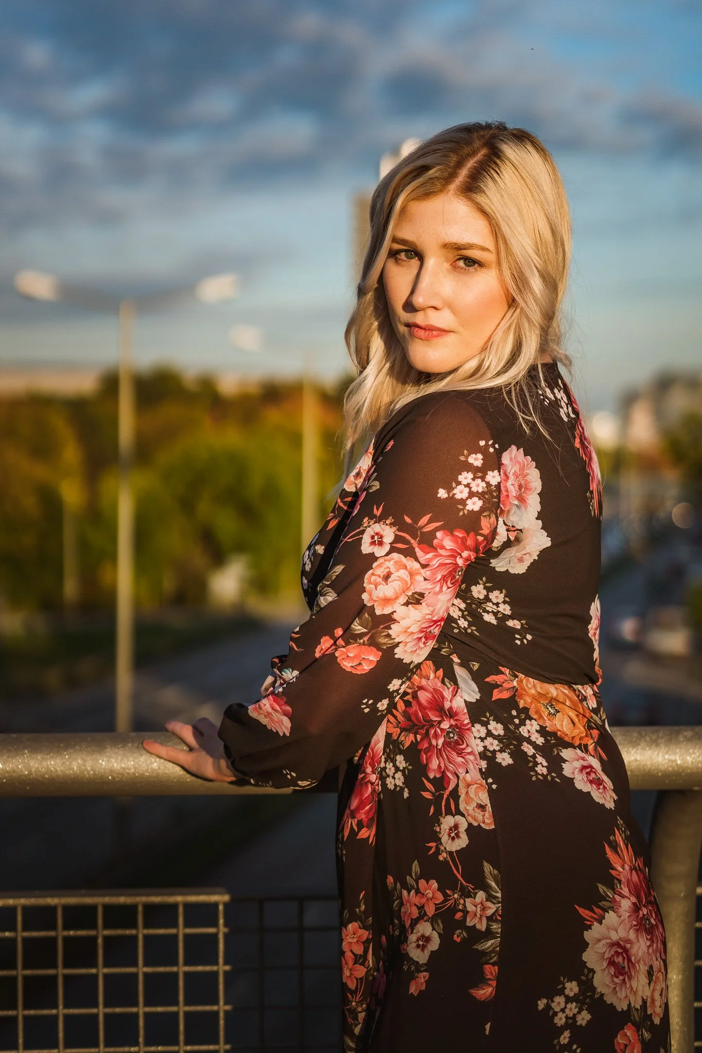 Eine junge blonde Frau in einem schwarzen Kleid mit rosa und weißen Blumenmustern steht an einer Brüstung und blickt in die Kamera, im Hintergrund ist ein bewölkter Himmel und eine Straße zu sehen.