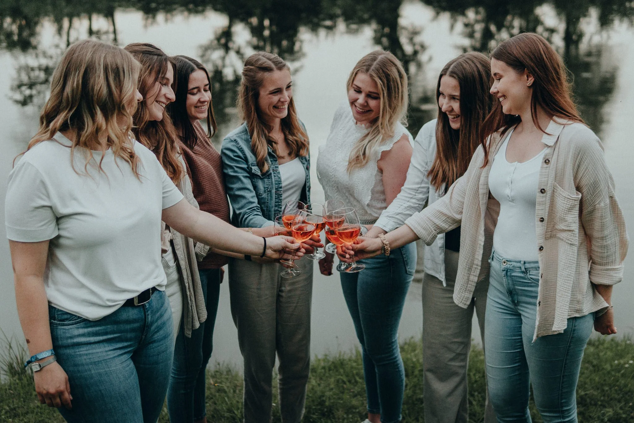 Sieben Frauen stoßen mit Roséwein in einem Kreis am Wasser an.