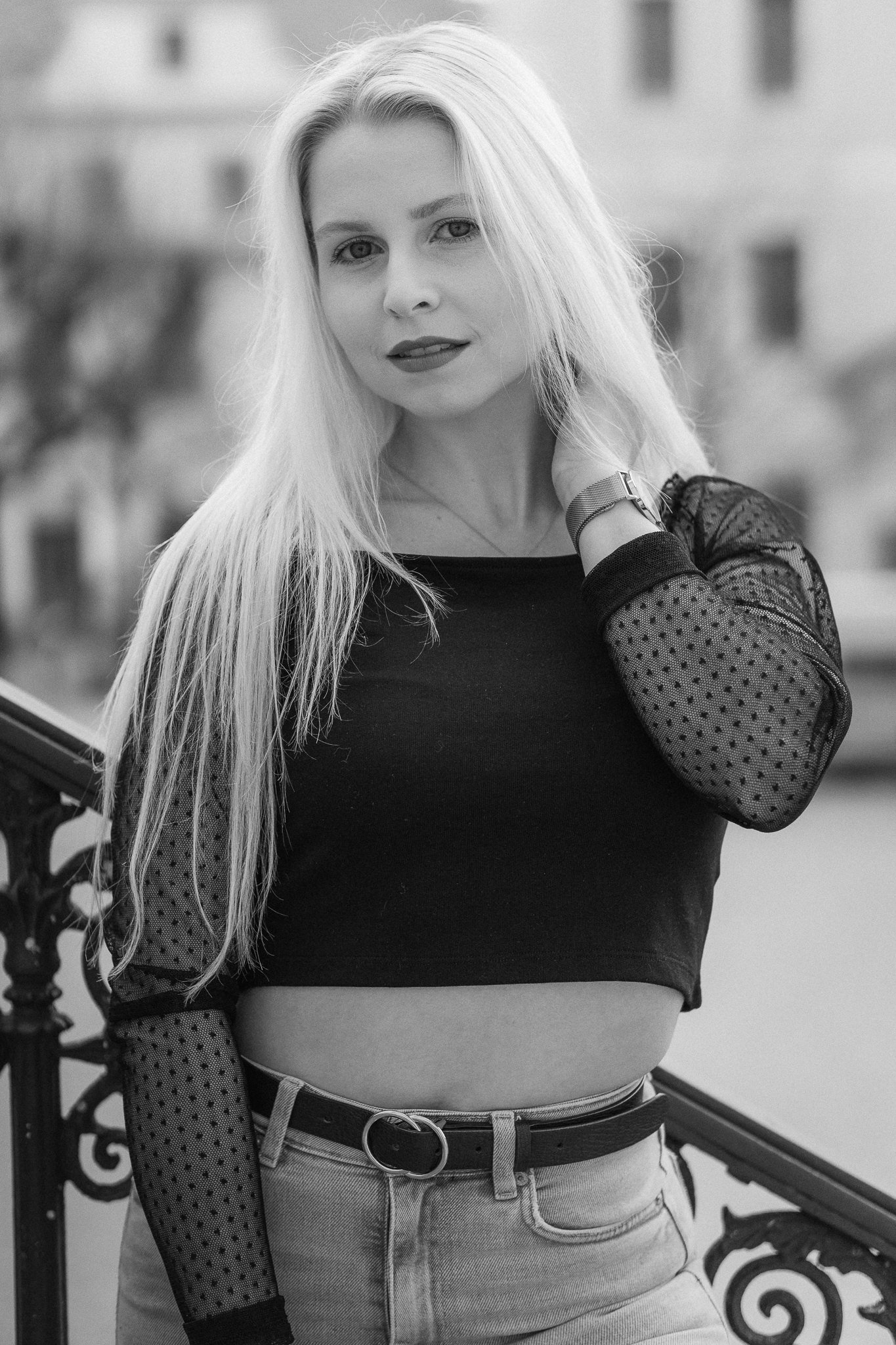 Schwarz-Weiß-Foto einer jungen Frau mit langen Haaren, die an einem Geländer steht, lächelnd in die Kamera blickt, trägt ein schwarzes Oberteil mit transparenten, gepunkteten Ärmeln, eine Armbanduhr und eine Jeans