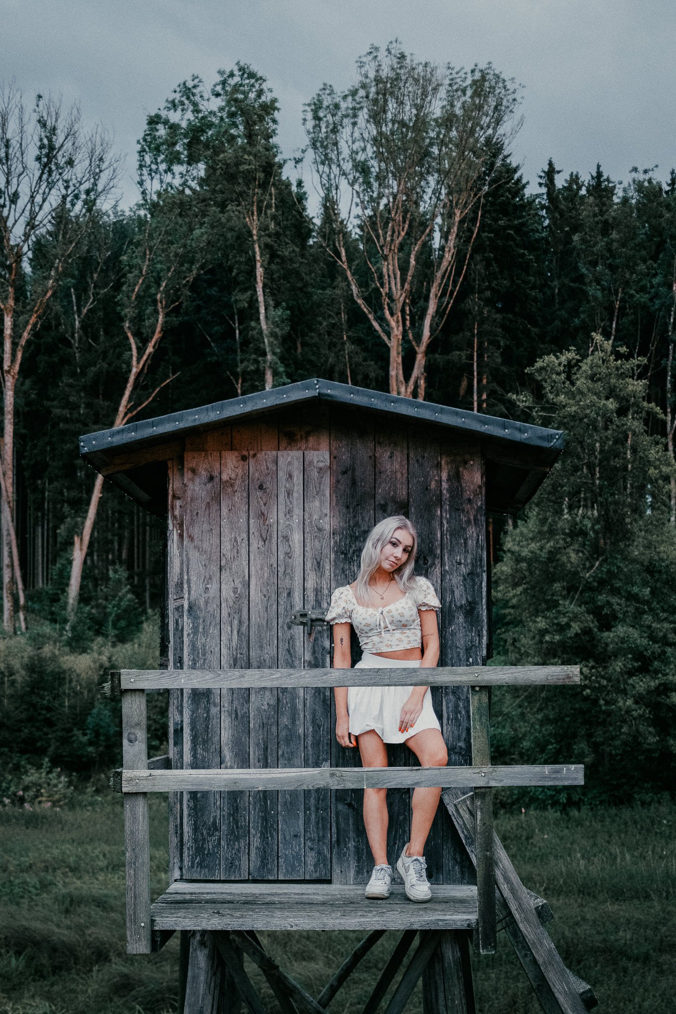 Junge Frau steht auf einer kleinen Holzbaustelle in einem Wald, trägt ein weißes schulterfreies Oberteil und einen weißen Rock, bei Dämmerlicht