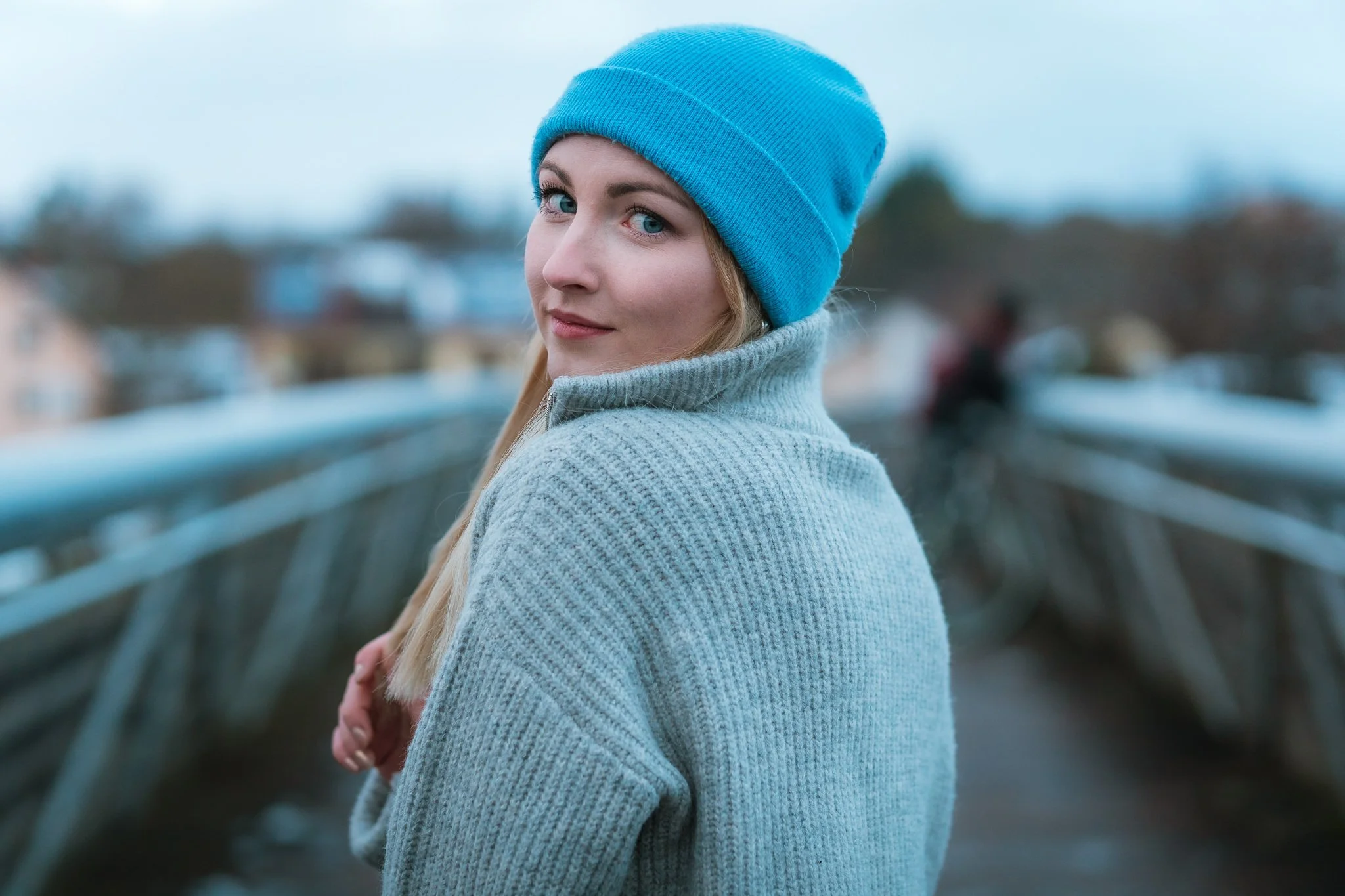 Junge Frau trägt blaue Mütze und grauen Pullover auf einer Brücke, blickt zurück, bei bewölktem Himmel im Freien.