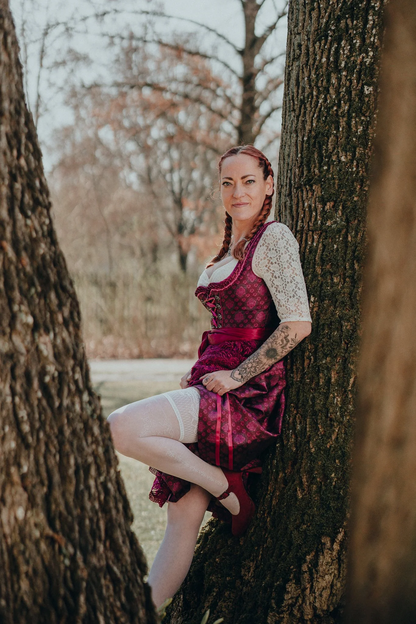 Eine Frau in traditioneller bayerischer Tracht sitzt an einer Baumstamm im Freien, umgeben von Bäumen ohne Blätter bei bewölktem Himmel.