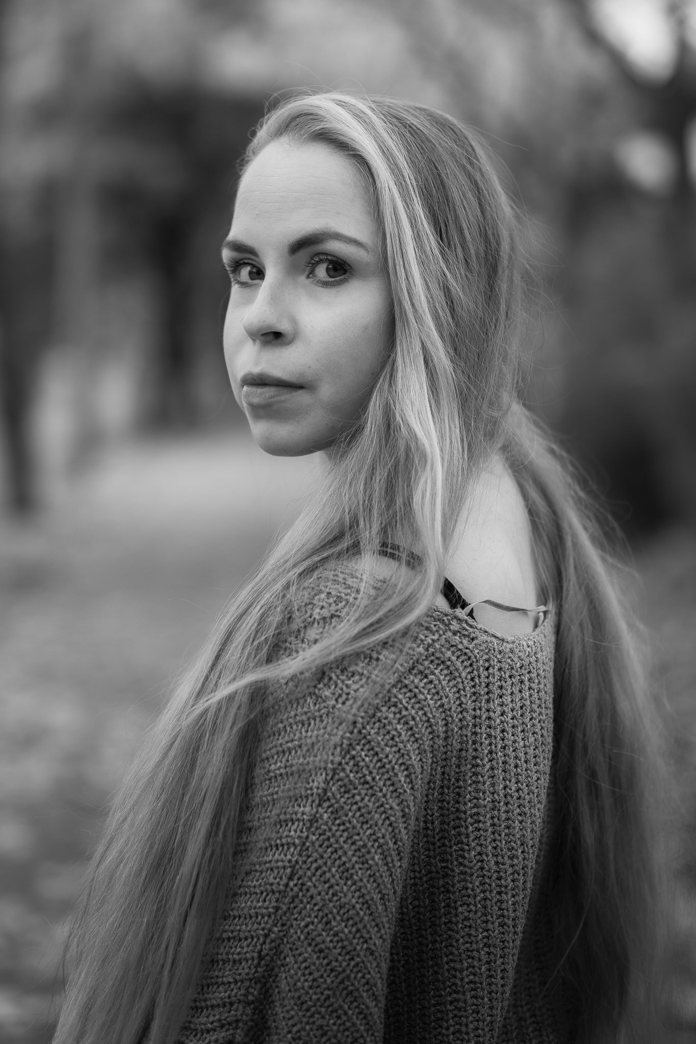 Schwarz-Weiß-Fotografie einer jungen Frau mit langem, gewelltem Haar, die nach rechts schaut, im Freien mit unscharfem Hintergrund von Bäumen.