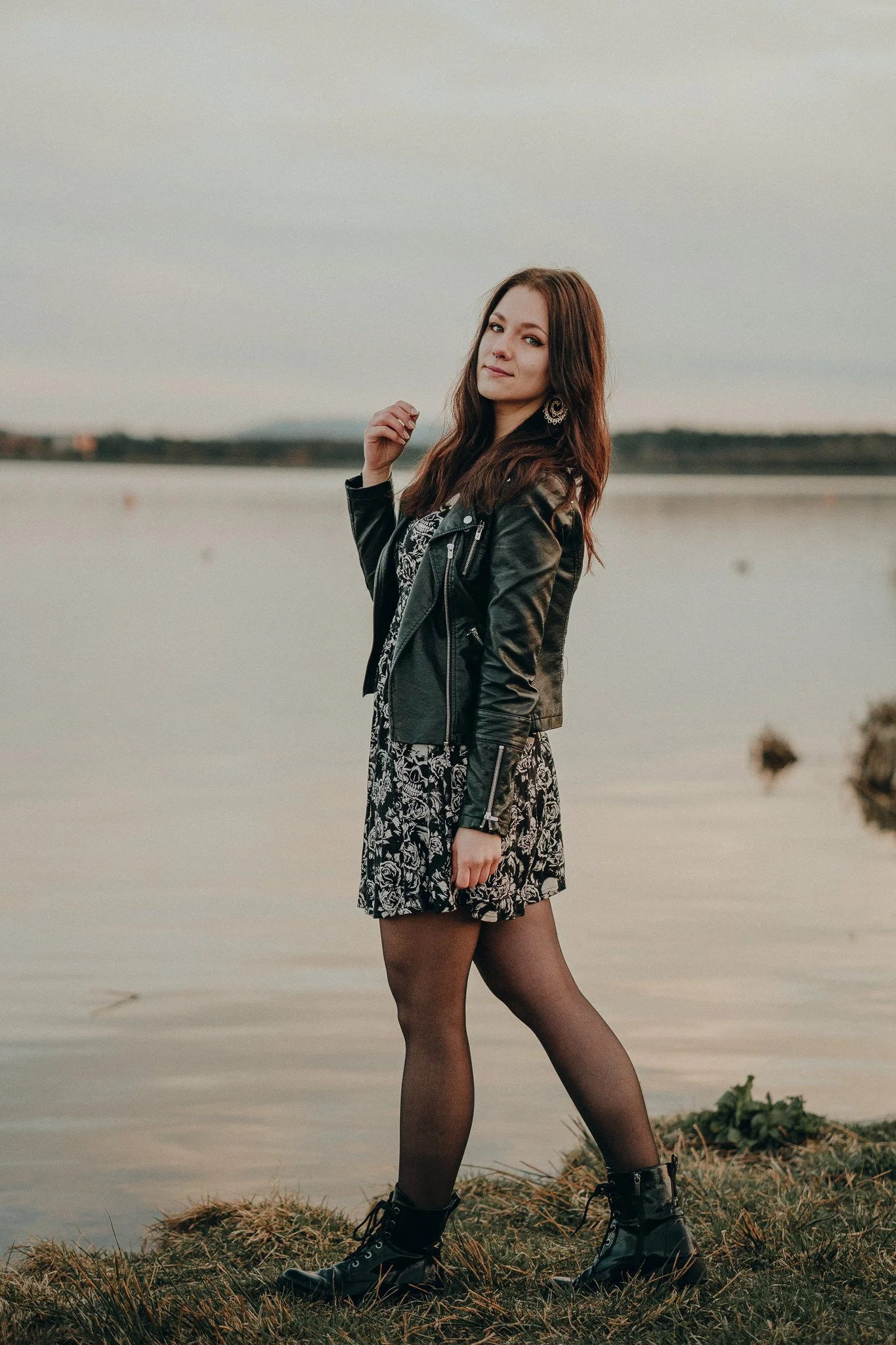 Junge Frau mit braunen Haaren steht an einem See, trägt ein schwarzes Lederjacke, ein schwarzes Kleid mit Weißem Muster, schwarze Strümpfe und schwarze Stiefel, bei Sonnenuntergang.