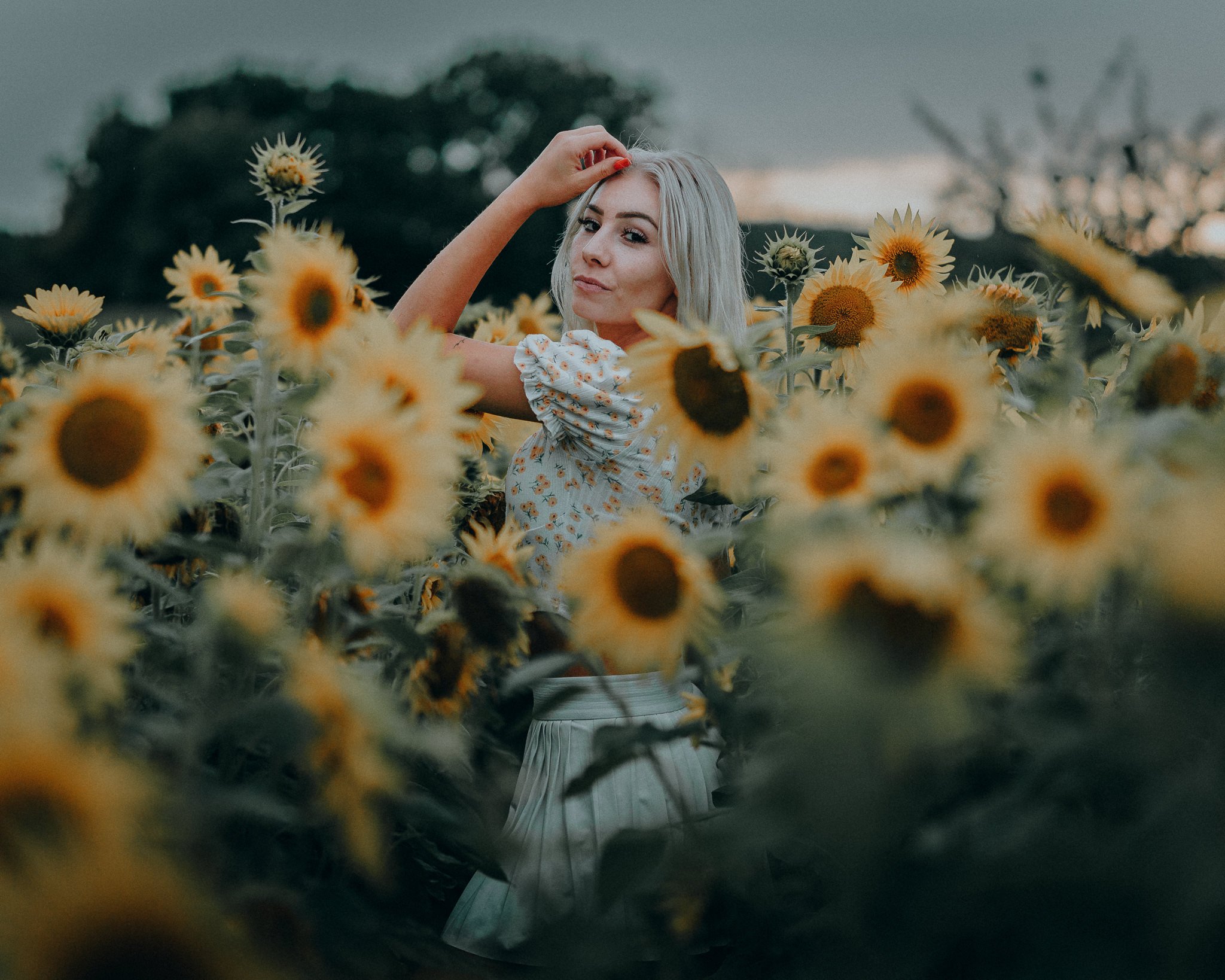 Eine junge Frau steht in einem Sonnenblumenfeld bei Sonnenuntergang, trägt ein geblümtes Oberteil und einen weißen Rock, schaut in die Kamera und hebt eine Hand an den Kopf.
