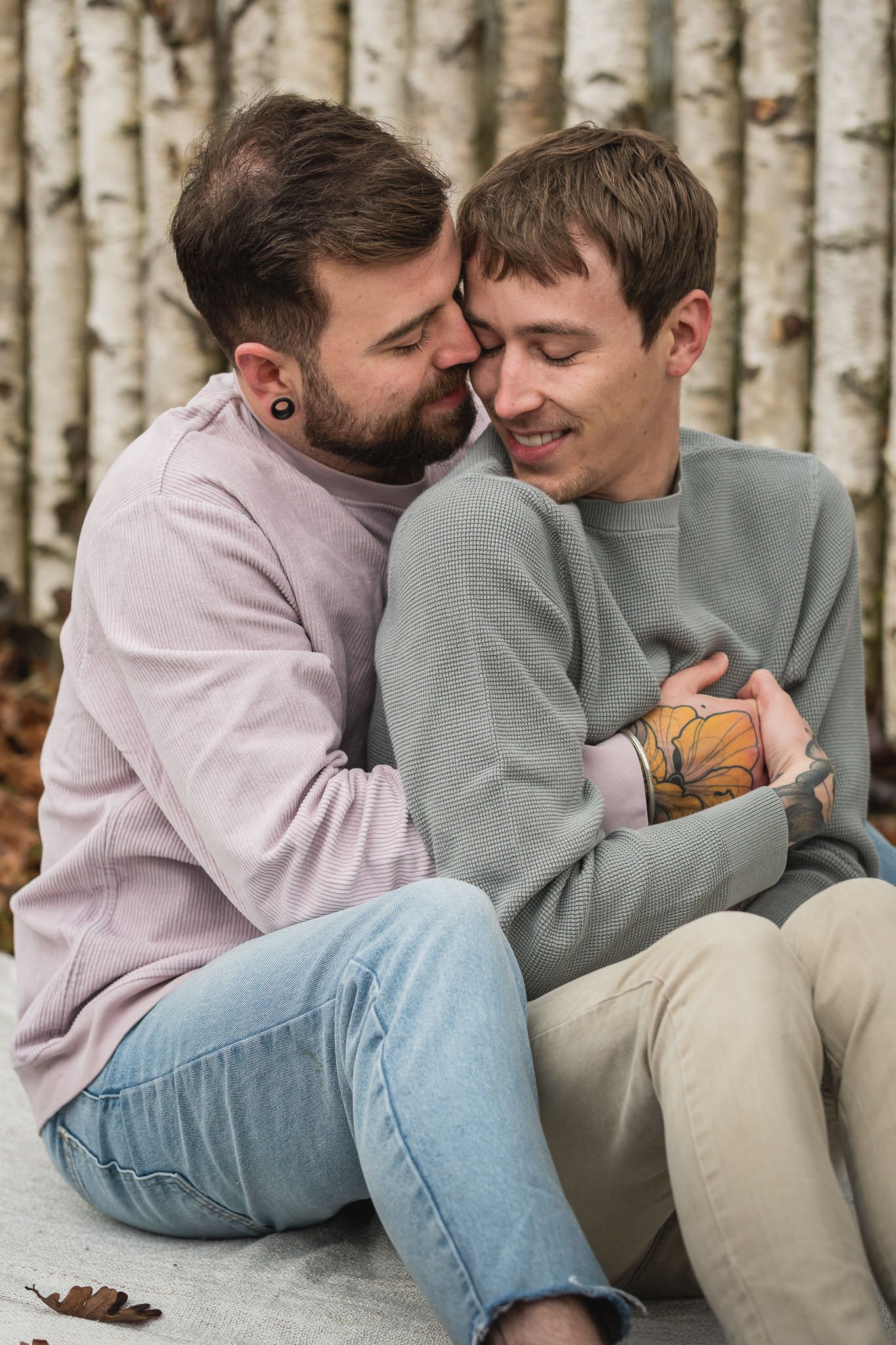 Zwei Männer sitzen eng umarmt vor einem Baumstamm im Freien, lächeln und drücken ihre Stirn aneinander.