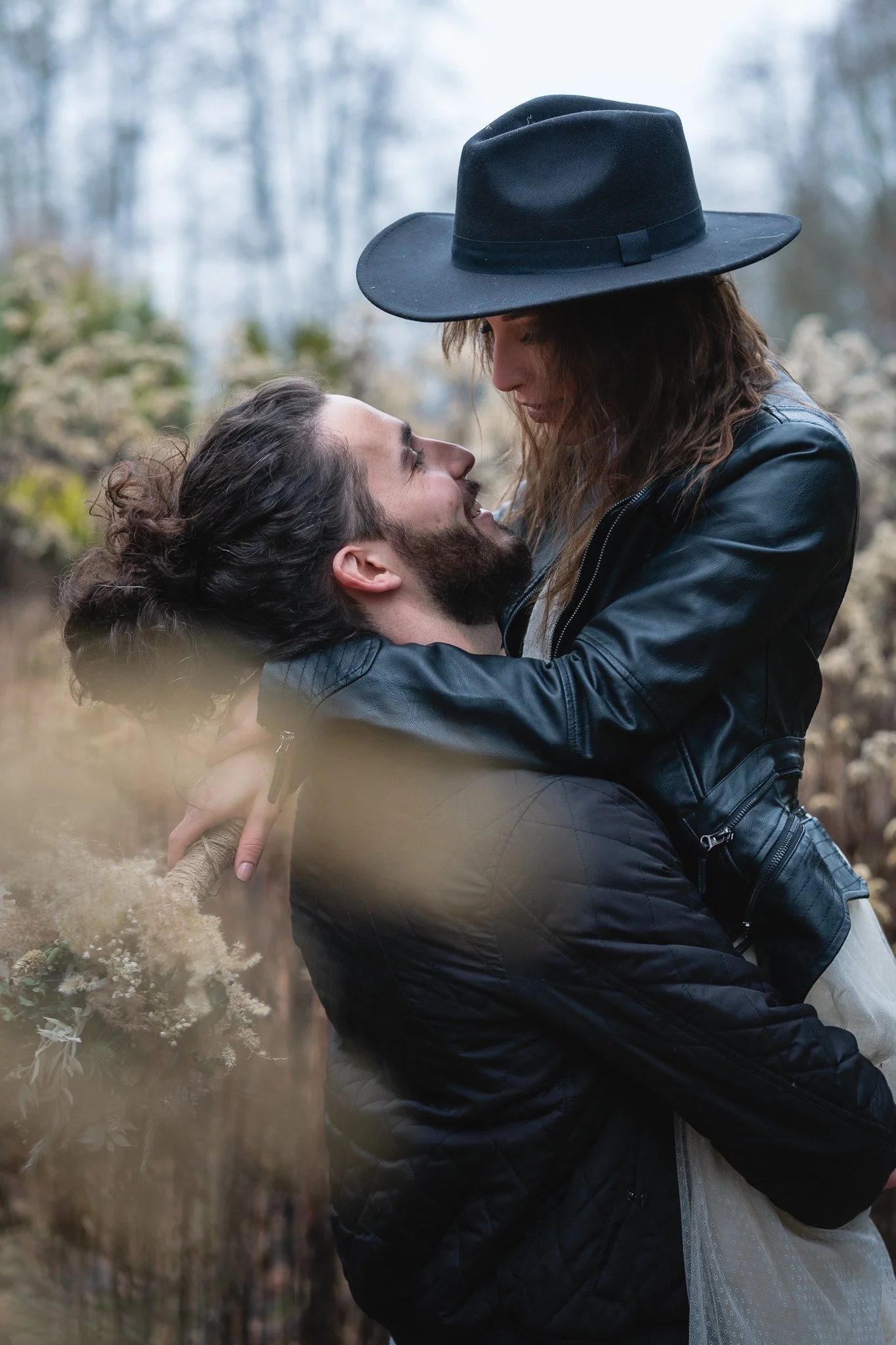 Ein Mann mit langen, lockigen Haaren und Bart hält eine Frau mit langen Haaren und Hut hoch umarmt, während sie sich ansehen, draußen im Freien.
