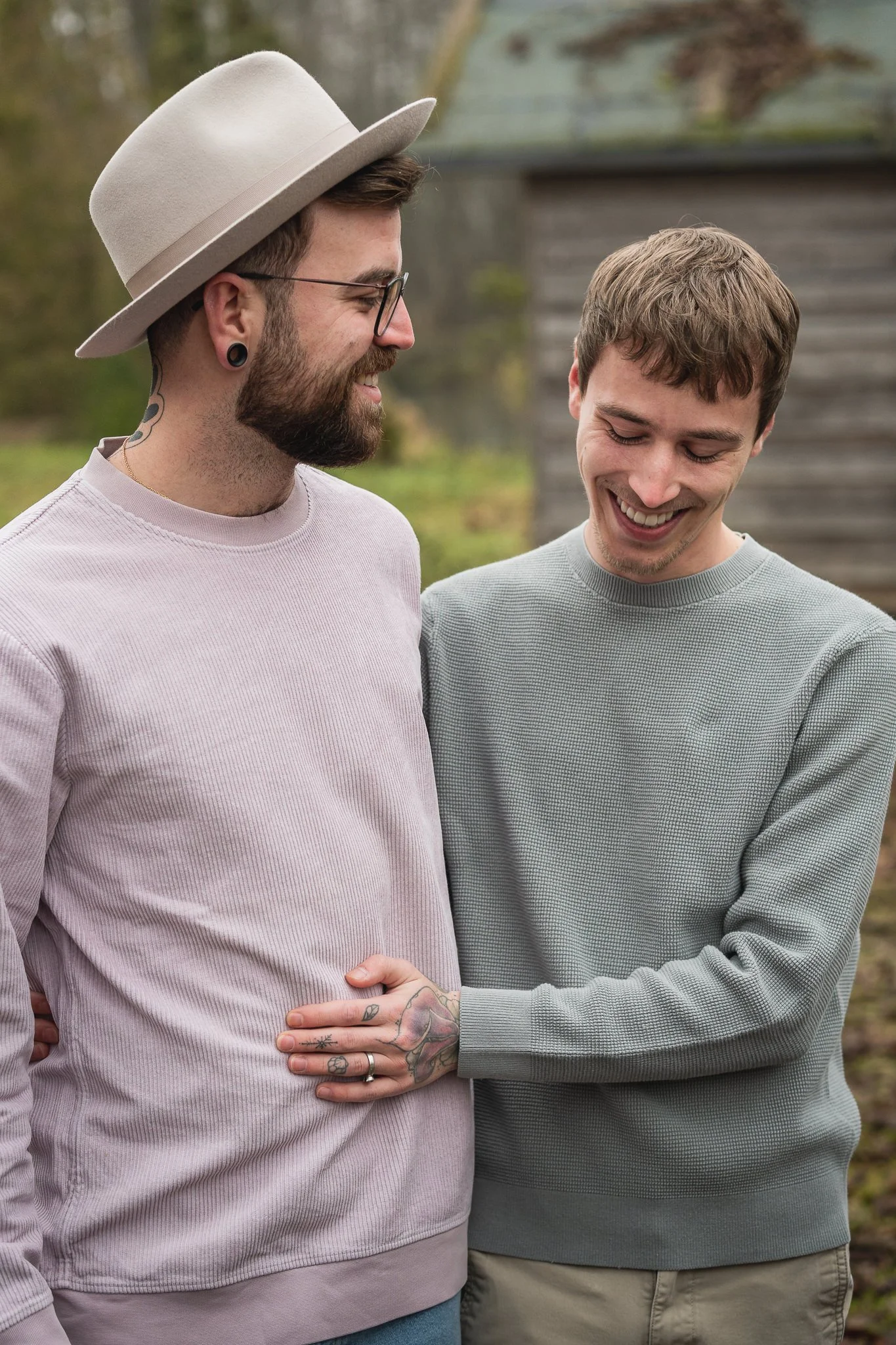 Zwei Männer lachen und umarmen sich im Freien, im Hintergrund ein altes Holzhaus.