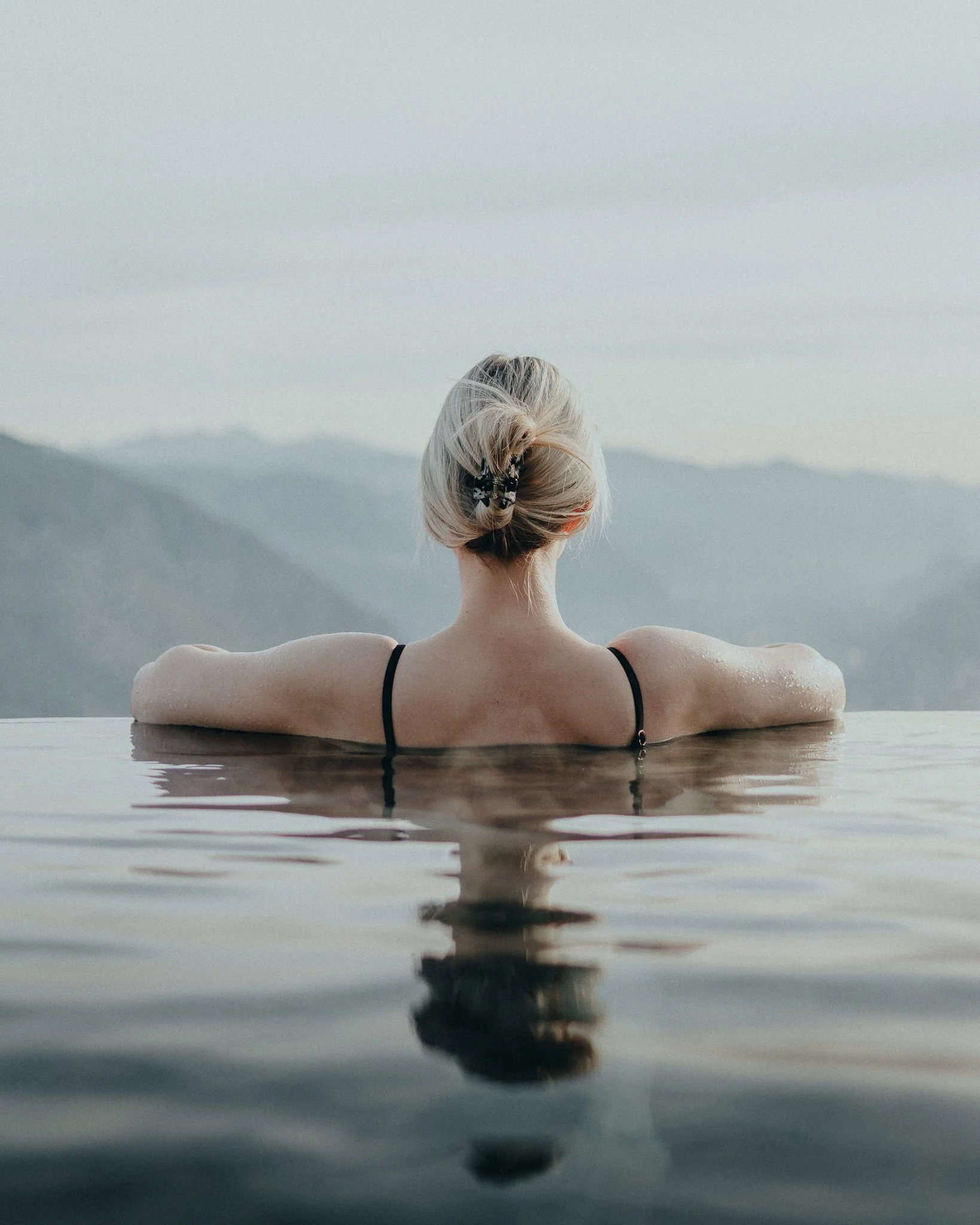 Frau mit blondem Haar in einem schwarzen Bikini in einem Infinity-Pool mit Bergkulisse im Hintergrund.