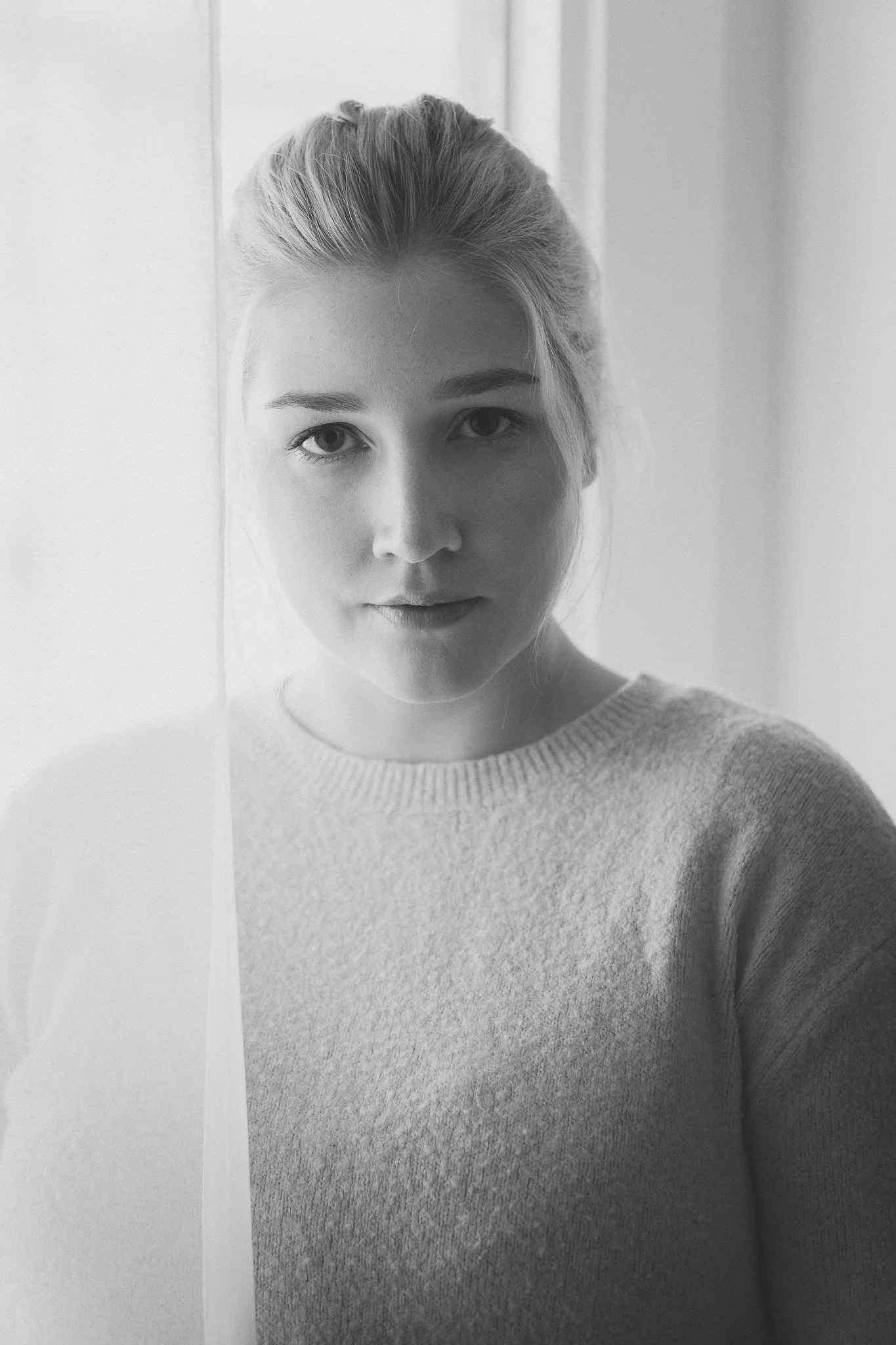 Schwarz-weiß Porträt eines jungen Mädchens mit hellen Haaren, die in einem hellen Raum neben einer Tür steht, Blick direkt in die Kamera.