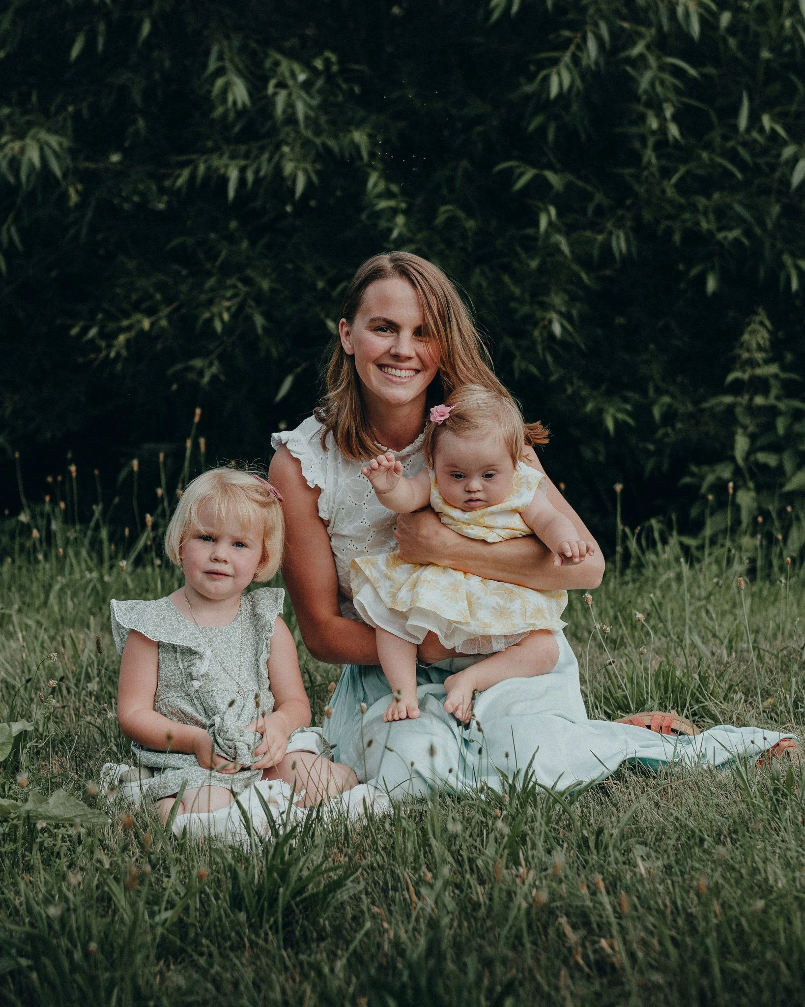 Eine Frau mit zwei kleinen Mädchen sitzt auf dem Gras im Freien, umgeben von Büschen und Blumen.