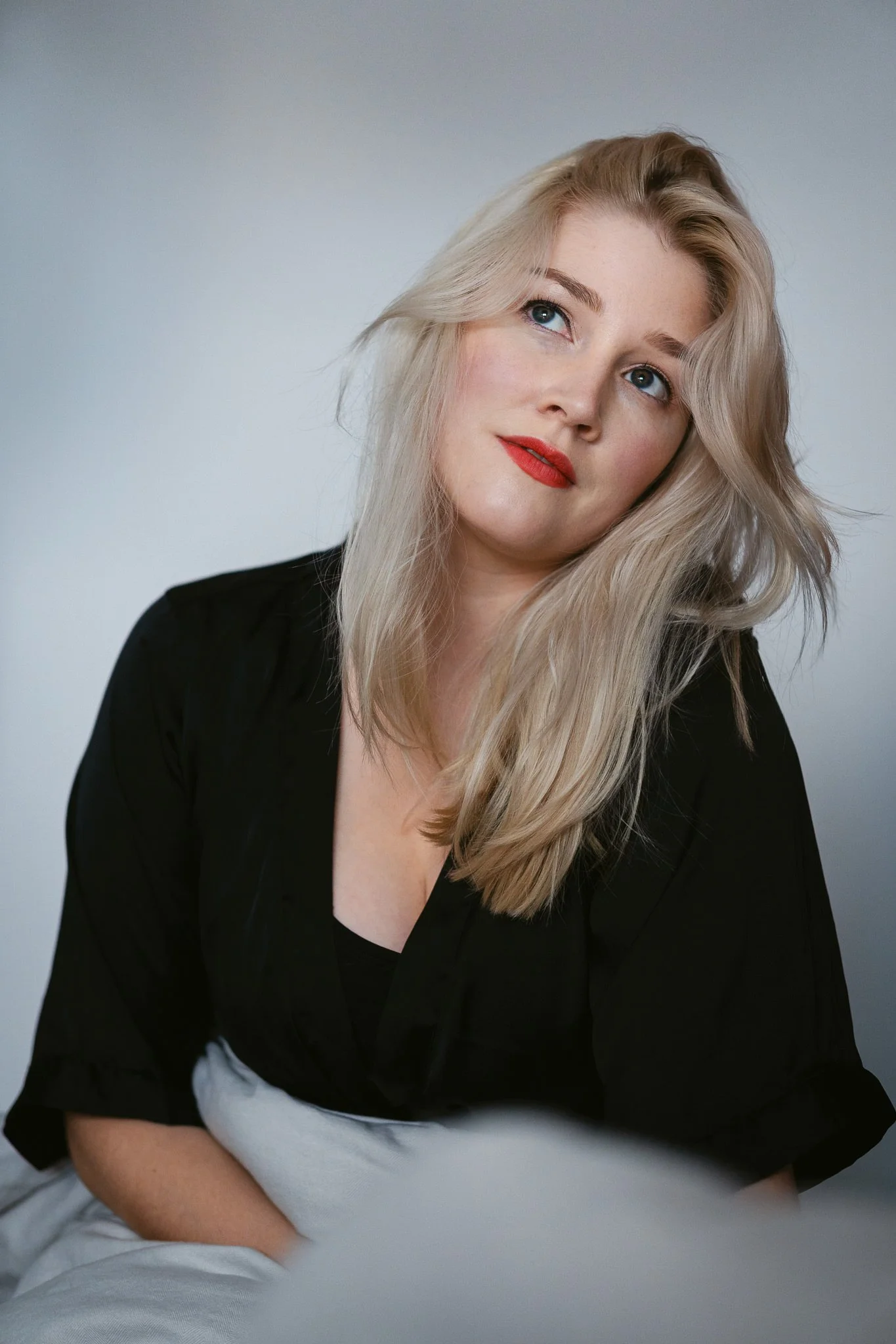Junge blonde Frau mit rotem Lippenstift schaut leicht nach oben, trägt schwarze Bluse, sitzt auf einem weißen Stuhl, heller Hintergrund.
