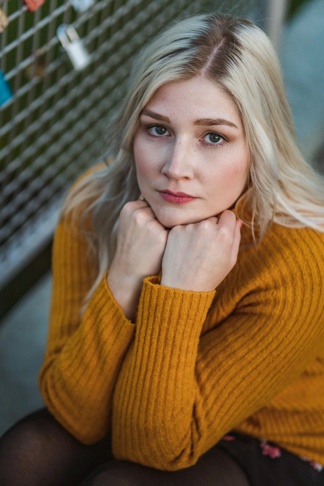 Ein junges blondes Mädchen sitzt mitunter einem Rost und schaut in die Kamera, das Kinn auf die Hand gestützt, trägt einen gelben Pullover.