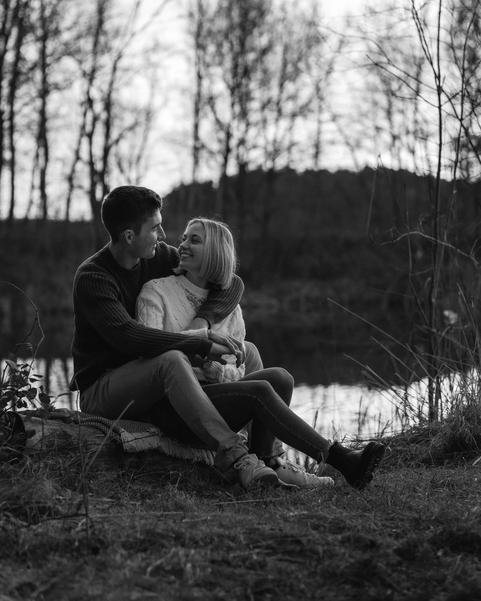Ein Paar sitzt am Ufer eines Sees, umgeben von Bäumen, und lächelt sich beim Blick in die Augen an.