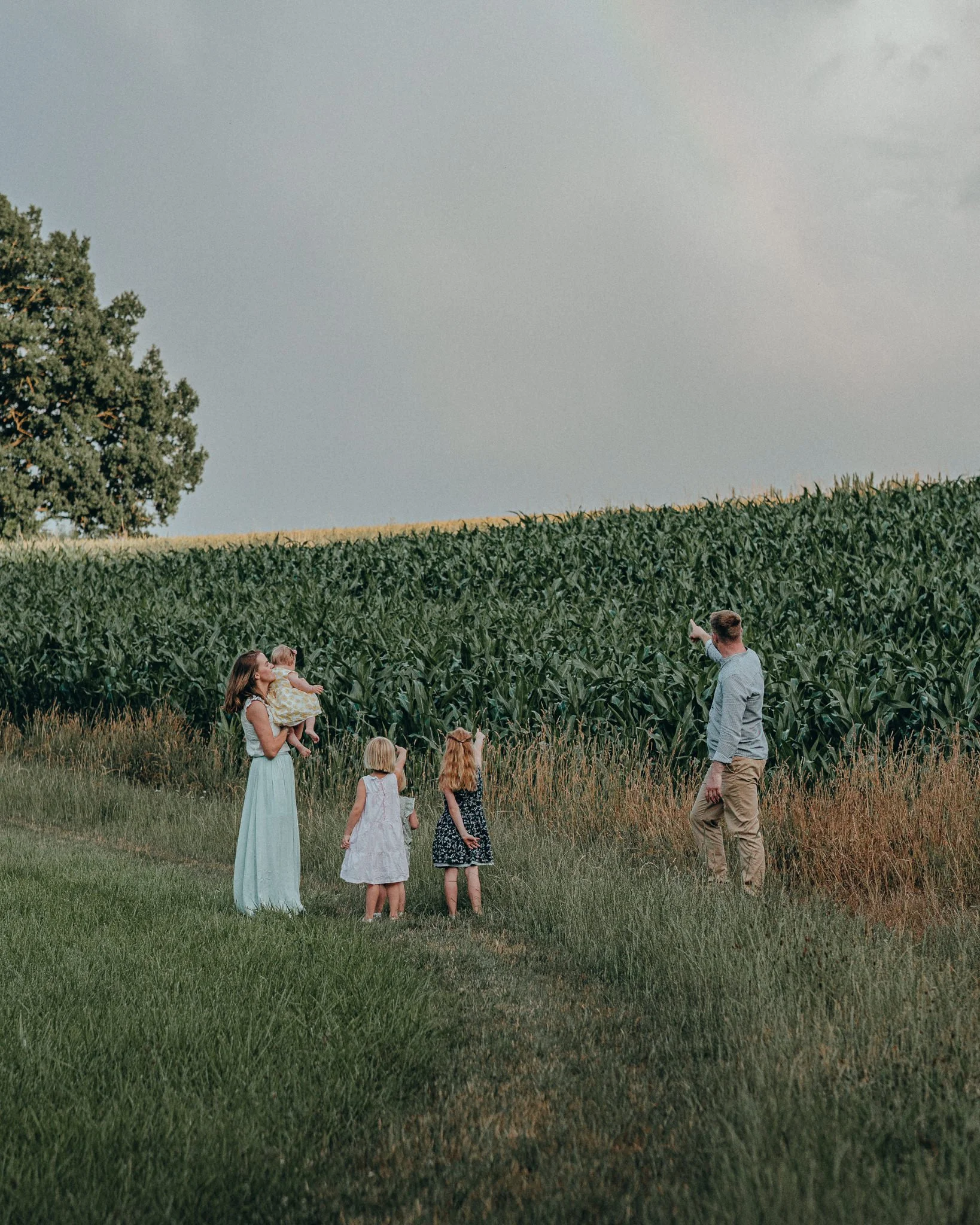 Familie mit Kindern im Grünen, der Vater zeigt auf eine Maispflanze im Feld, die Mutter hält ein kleines Kind im Arm.