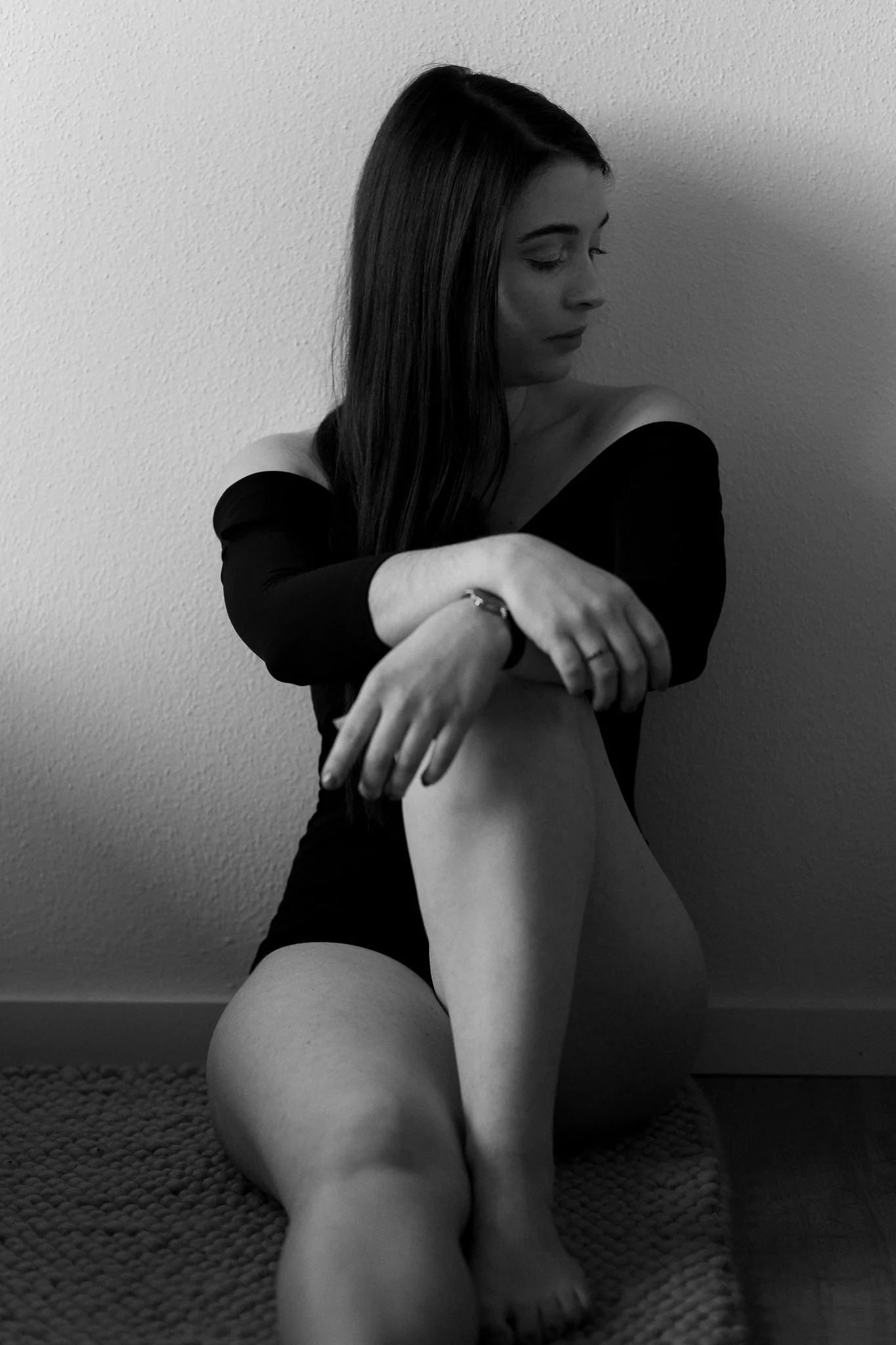 Schwarz-weiße Aufnahme einer jungen Frau, die auf dem Boden sitzt, die Beine anzieht und den Blick nach unten richtet, mit geschlossenem Blick und entspanntem Ausdruck.