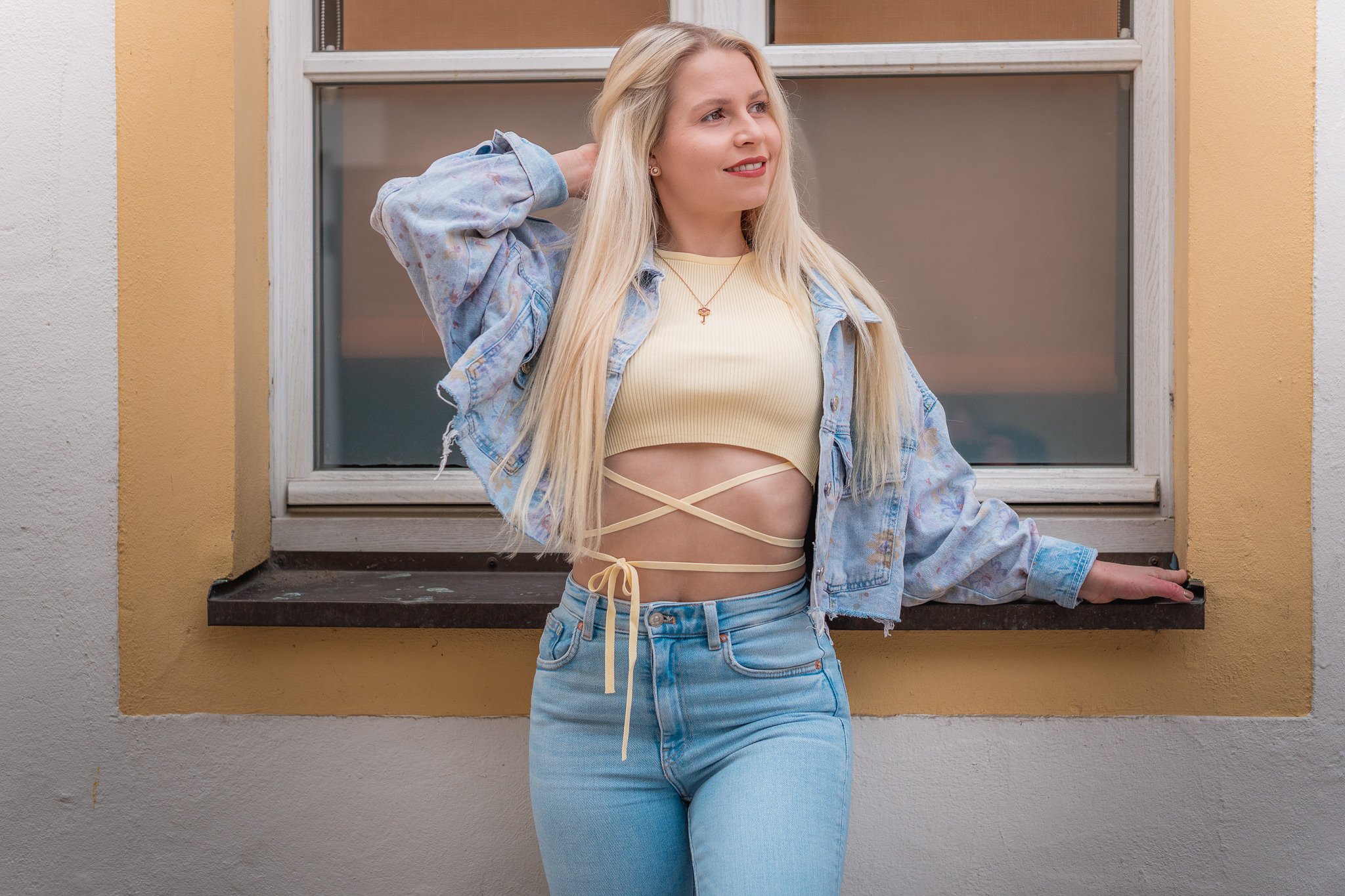 Junge Frau mit langem blondem Haar trägt eine gelbe Crop-Top, eine Jeansjacke, eine High-Waist-Jeans sowie eine Wickelcrossover-Kette vor einem Fenster an einer weißen und gelben Wand.