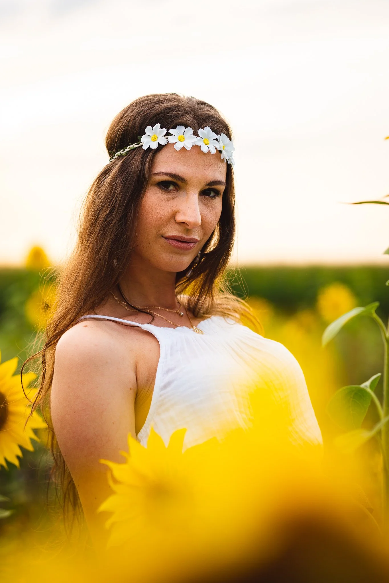 Junge Frau mit Blumenkranz am Kopf in einem Sonnenblumenfeld bei Sonnenuntergang.