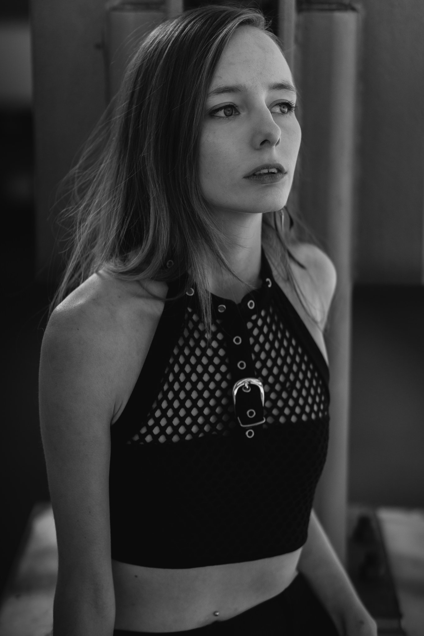 Schwarze und weiße Schwarz-Weiß-Fotografie eines jungen Mädchens mit mittellangem Haar, das auf einem Stuhl sitzt, trägt ein Netzoberteil und schaut nach rechts. Texturierte Wand im Hintergrund.