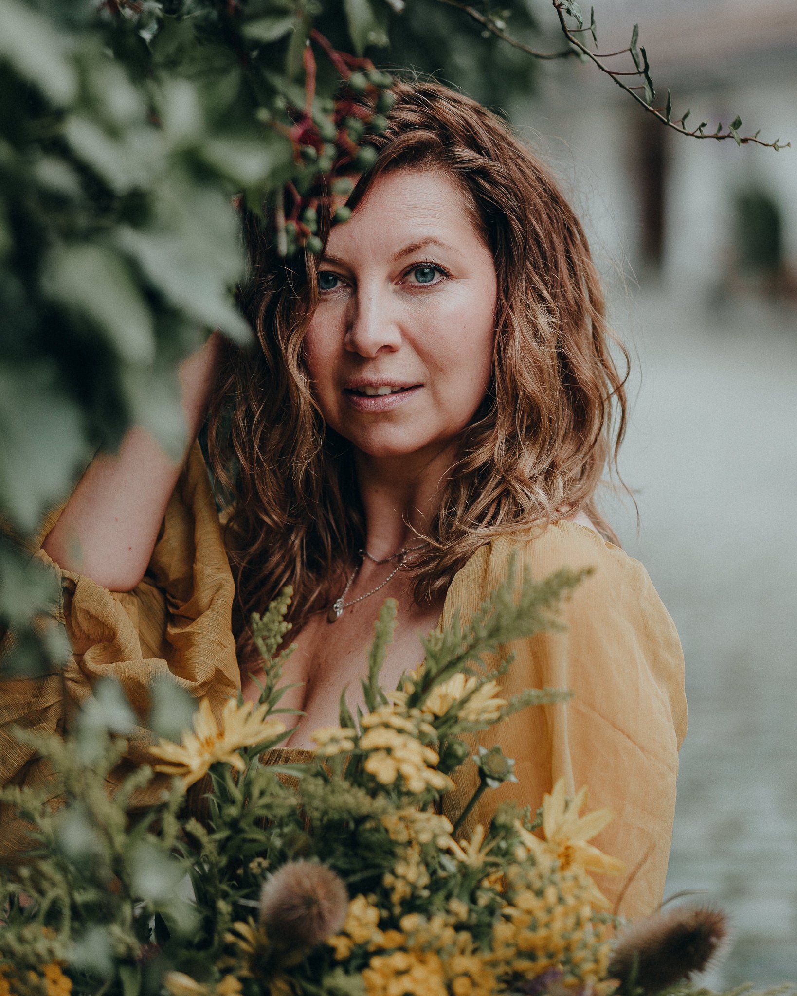 Eine Frau mit lockigem braunem Haar, die eine gelbe Bluse trägt, hält ein Blumenbouquet mit gelben und grünen Blumen vor einem Wasserhintergrund und schaut in die Kamera.
