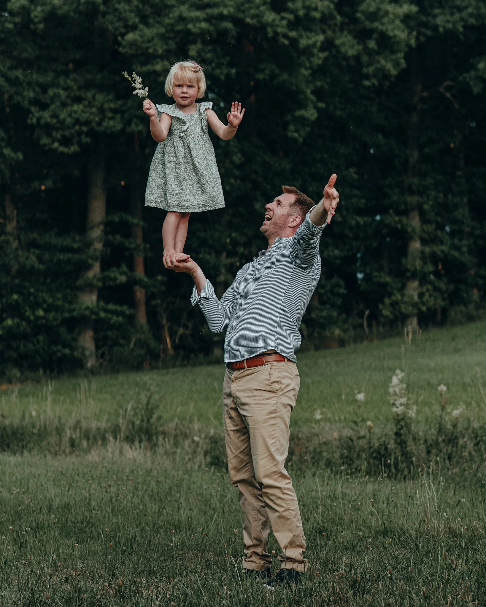Ein Mann hebt ein kleines Mädchen auf eine Hand, während sie im Freien auf einer grünen Wiese mit Bäumen im Hintergrund spielen.