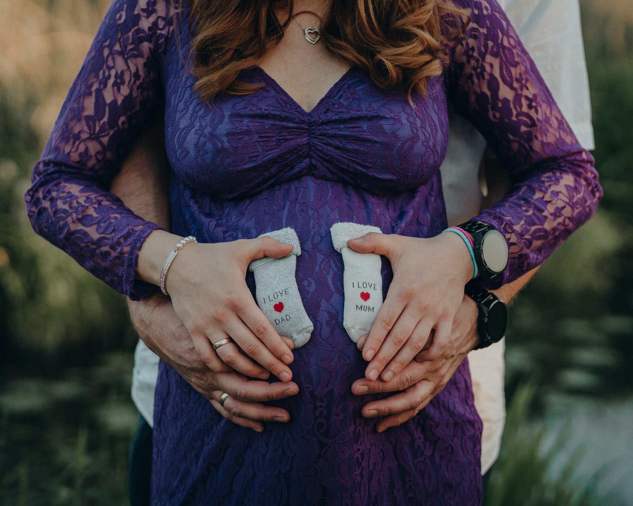Schwangere Frau in lila Kleid mit Schuhen, die 'I LOVE MUM' tragen, während ihr Partner sie liebevoll umarmt und ihre Babybauch hält.