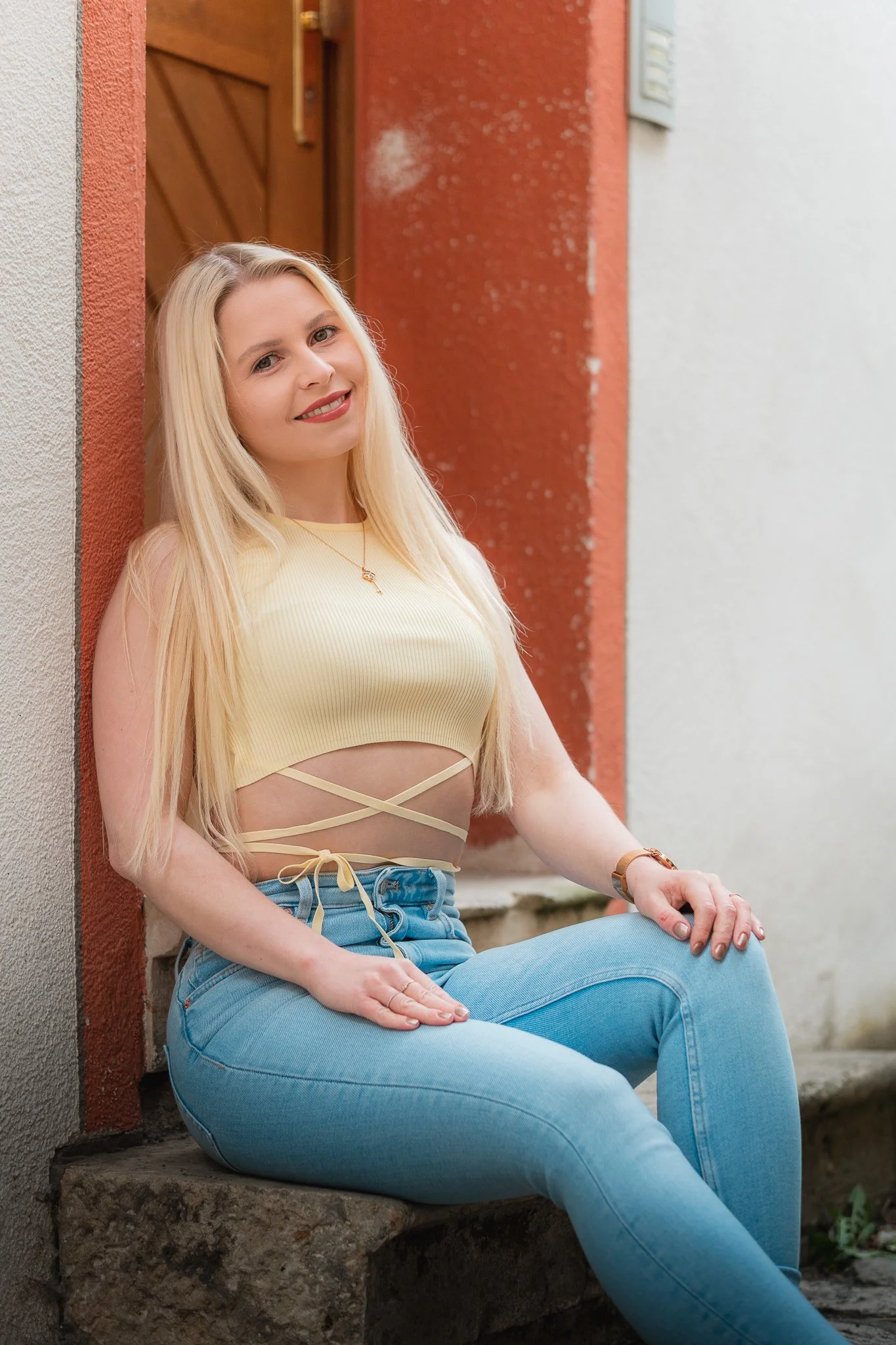 Junge Frau mit langen blonden Haaren, in einer hellgelben Top, sitzt auf einer Steinbank vor einer Hauswand und lächelt in die Kamera.