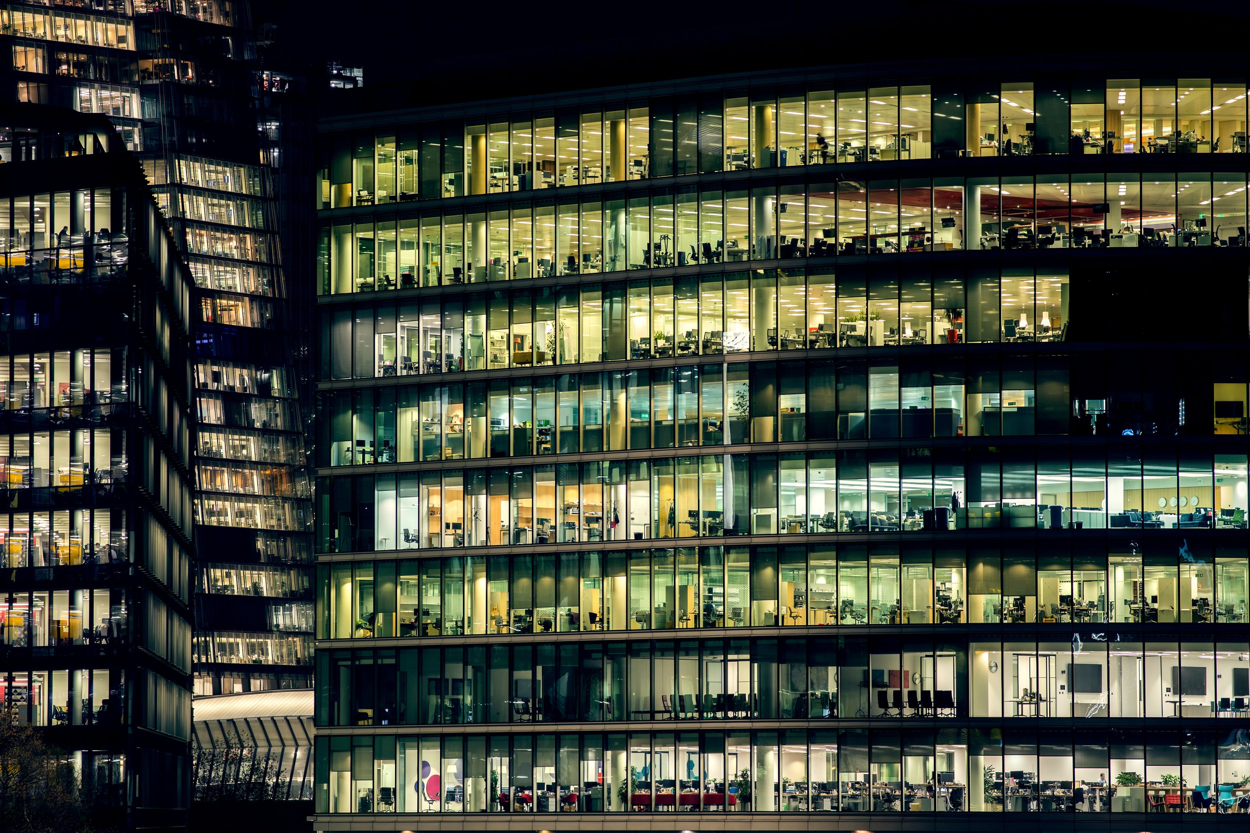Nighttime view of a modern office building with large glass windows, showing illuminated offices and workspaces on multiple floors.